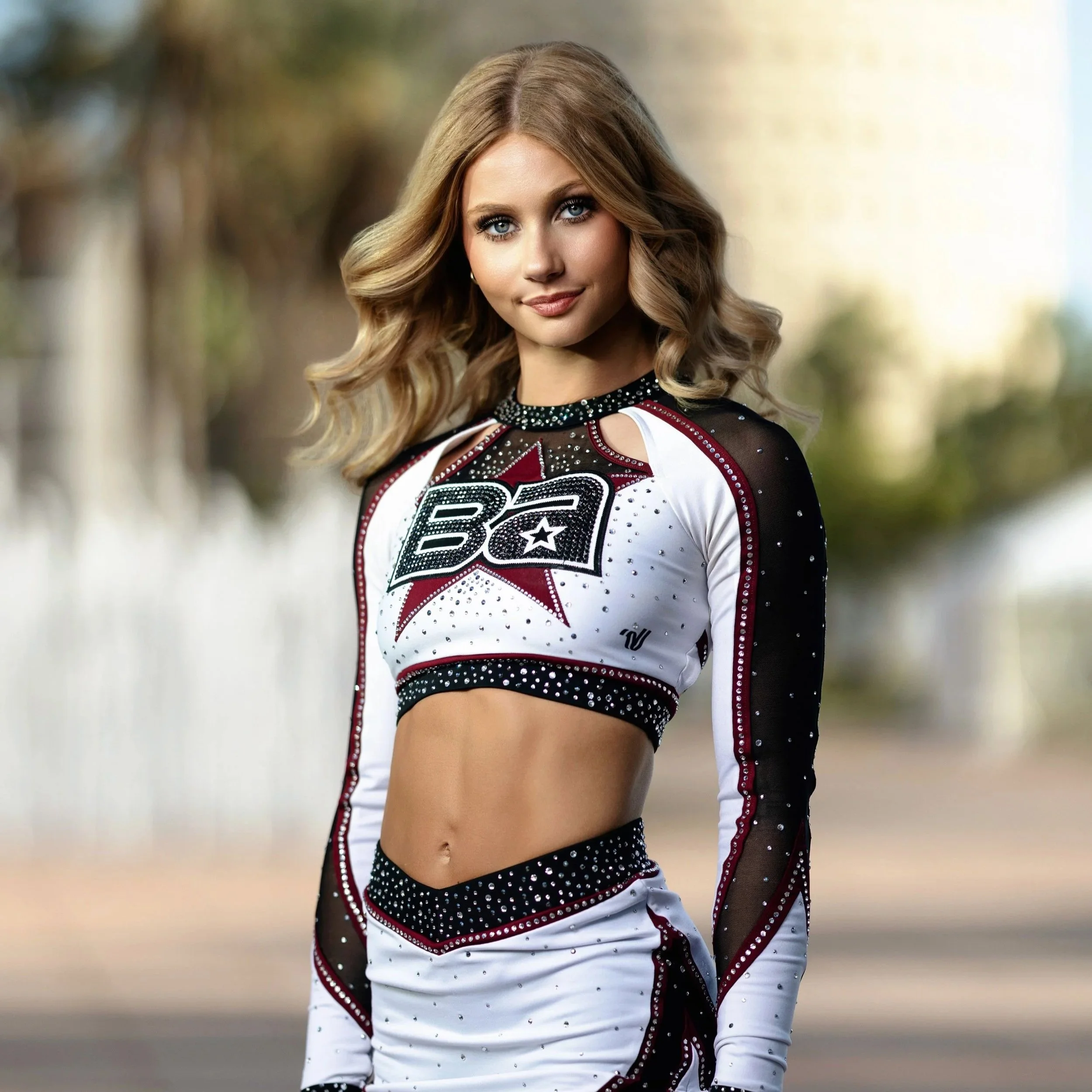 A young woman with long, wavy blonde hair and blue eyes, smiling slightly, wearing a white and black cheerleading outfit with red accents and rhinestones, posing outdoors.