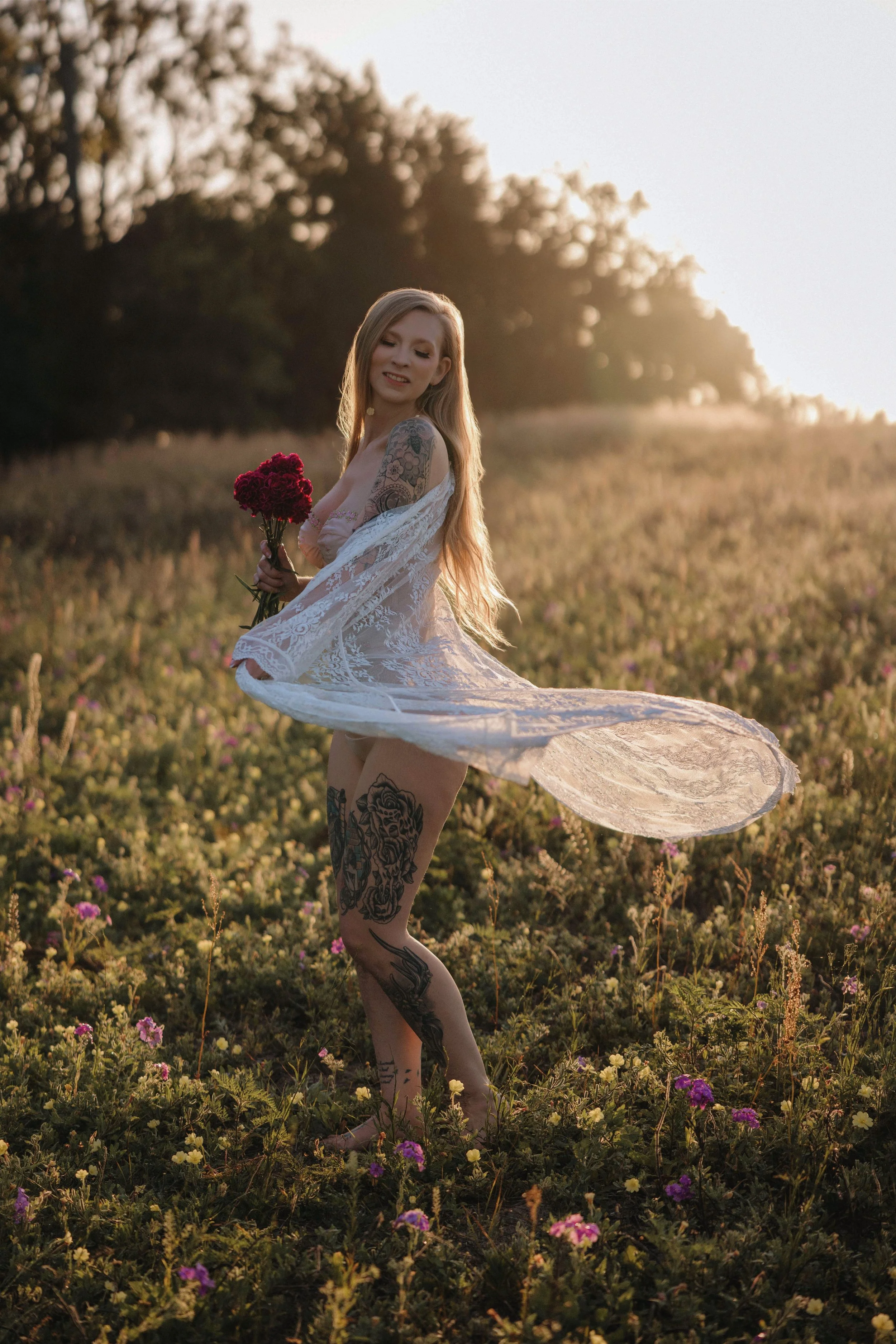 A woman with long blonde hair, tattoos on her legs and arms, wearing a white lace robe, standing in a field of yellow and purple flowers during sunset, holding a bouquet of red flowers, with trees in the background.