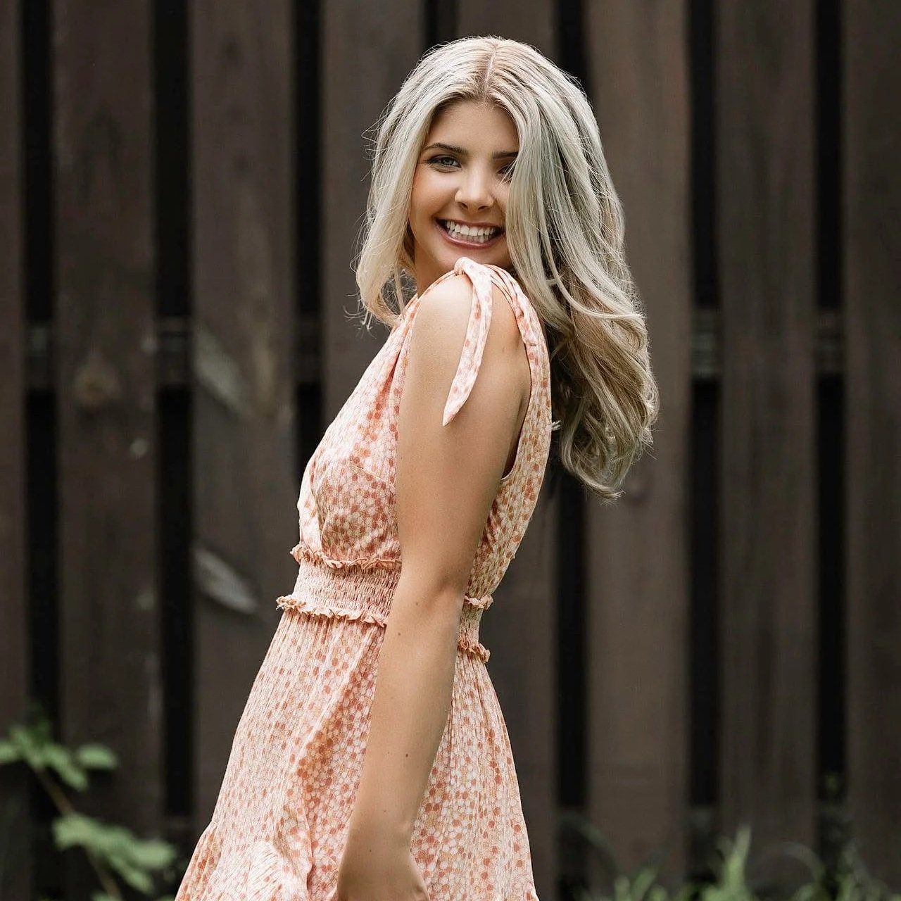 A young woman with long blonde wavy hair smiling and turning to her right against a wooden fence background.