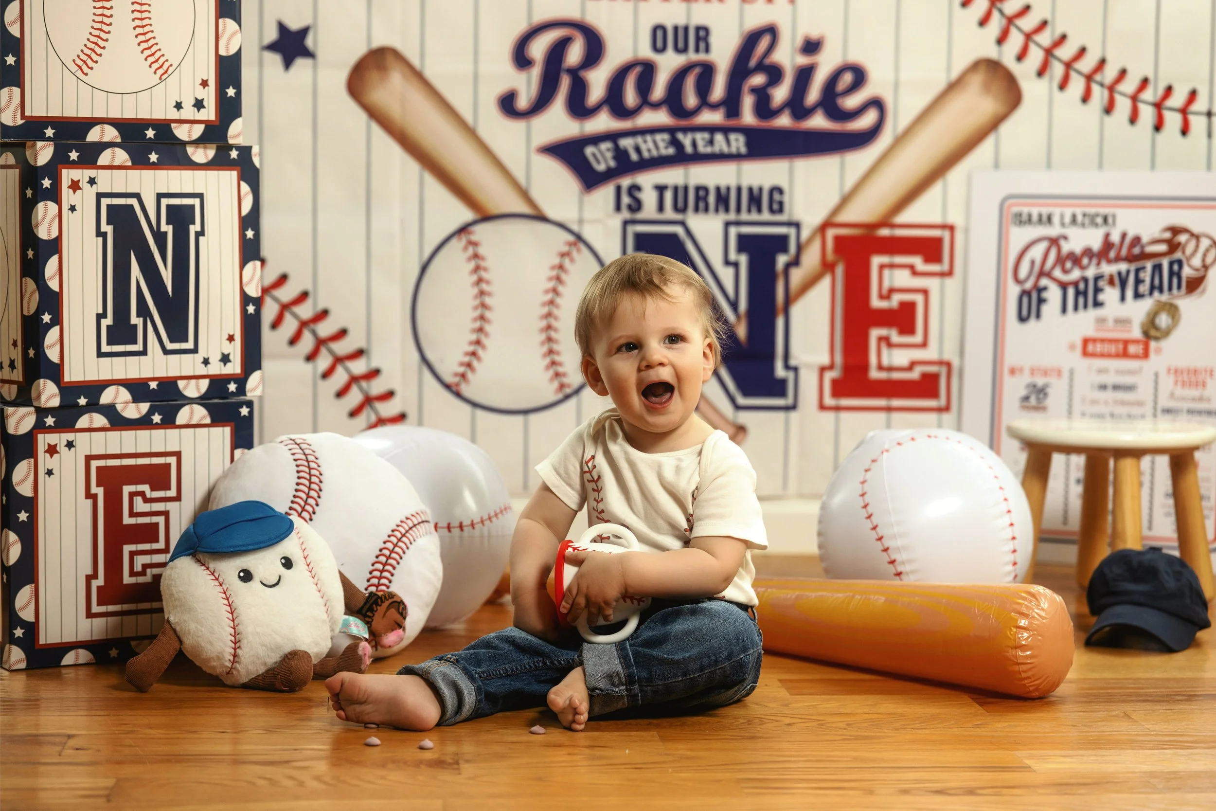 A young boy with blond hair sitting on the wooden floor surrounded by baseball-themed decorations, stuffed toys, and balloons in a room decorated for a baseball-themed celebration. The background features a large banner with the text 'Our Rookie of the Year is Turning' and large baseballs and baseball-themed posters.