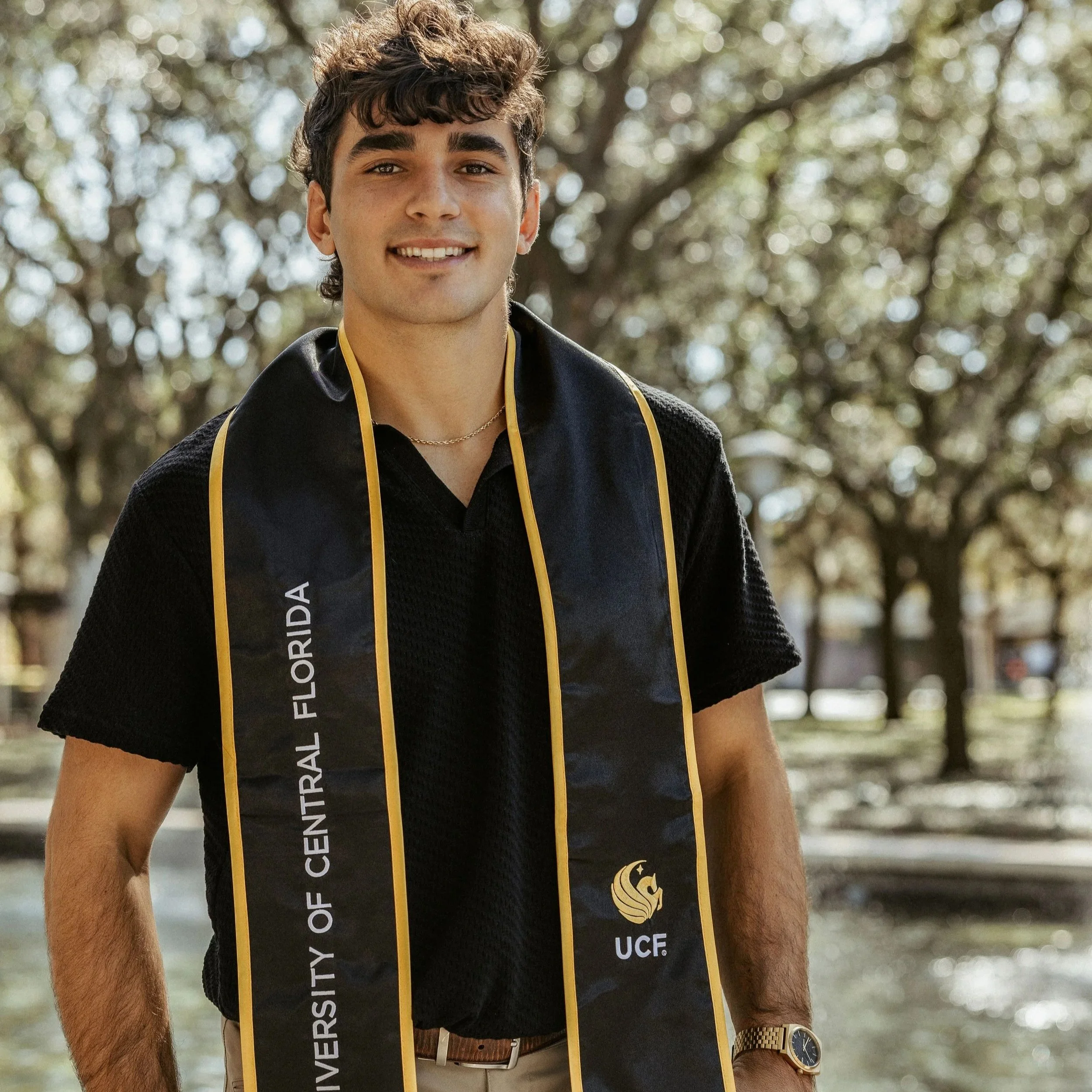 A young man with dark curly hair, smiling and wearing a black graduation gown with gold trim, stands outdoors with a background of trees and sunlight.