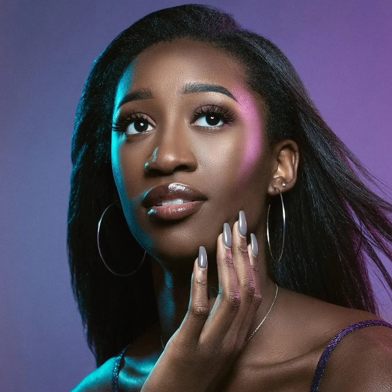 A close-up portrait of a Black woman with smooth skin, dark hair, and hoop earrings. She has makeup, including glossy lips, blush, and eye makeup. Her hand is near her face with styled nails, and the background has a gradient of purple and blue lighting.