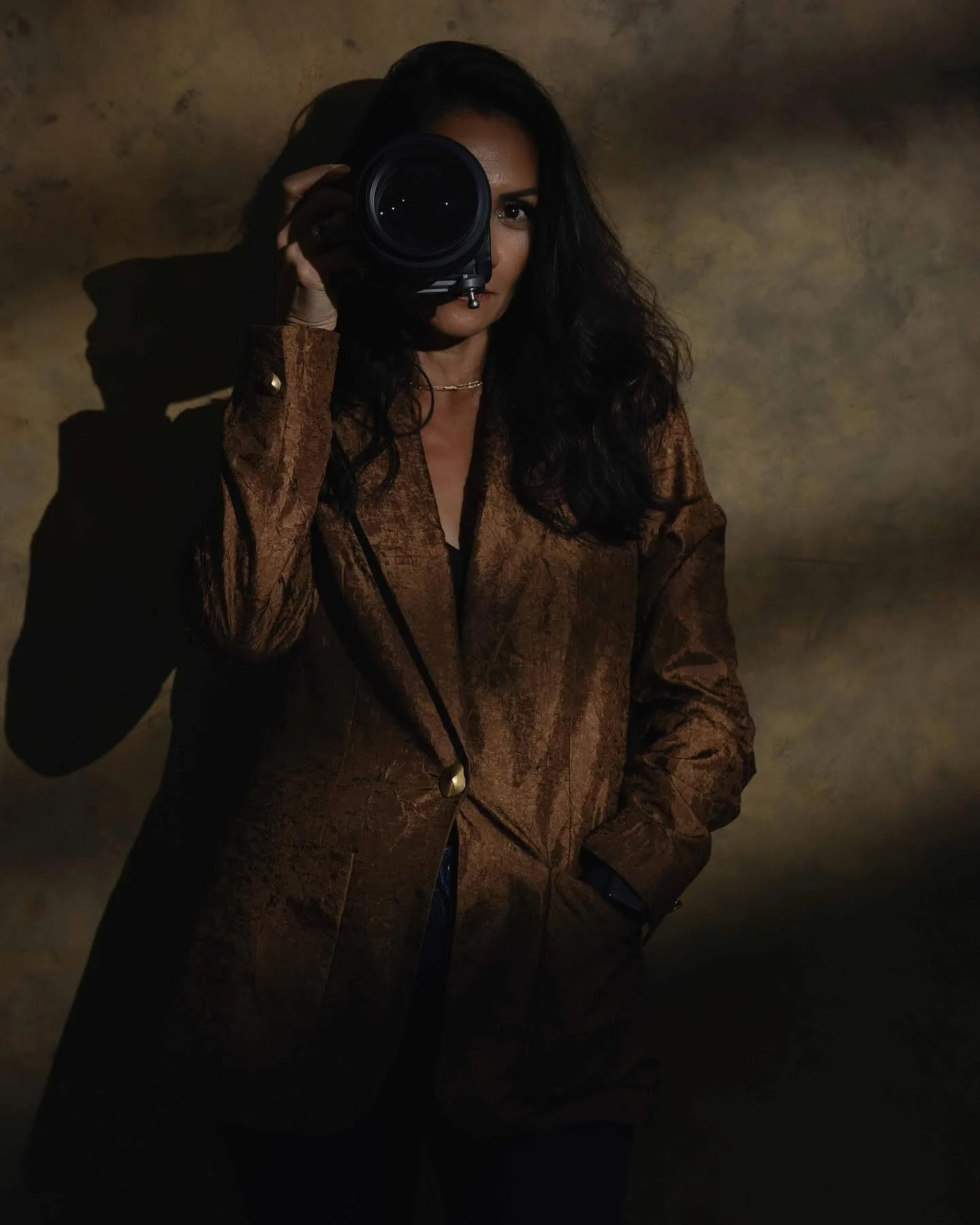 Woman with dark hair taking a photo with a camera, dressed in a brown leather jacket, against a brown textured background.