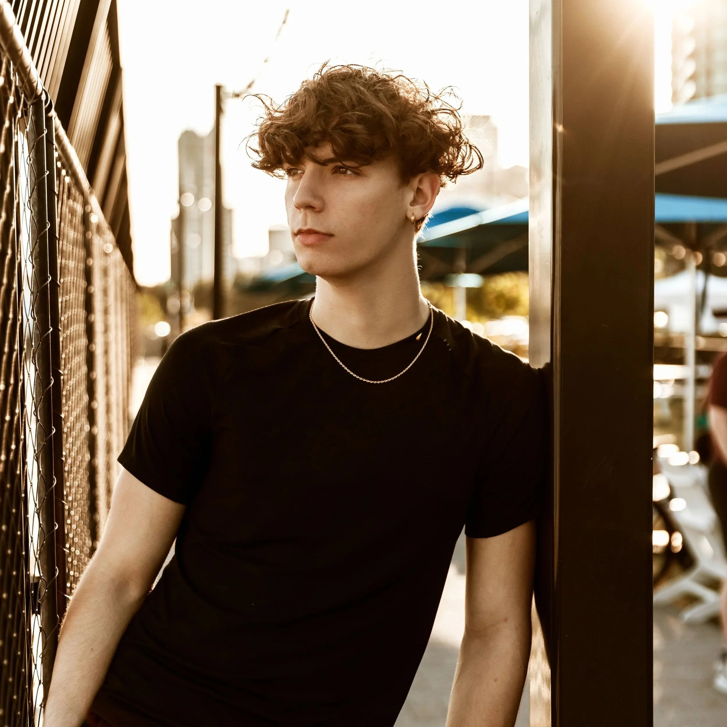 A young person with curly hair and earrings looking to the side, standing outdoors near a fence and a metal structure, with sunlight in the background.