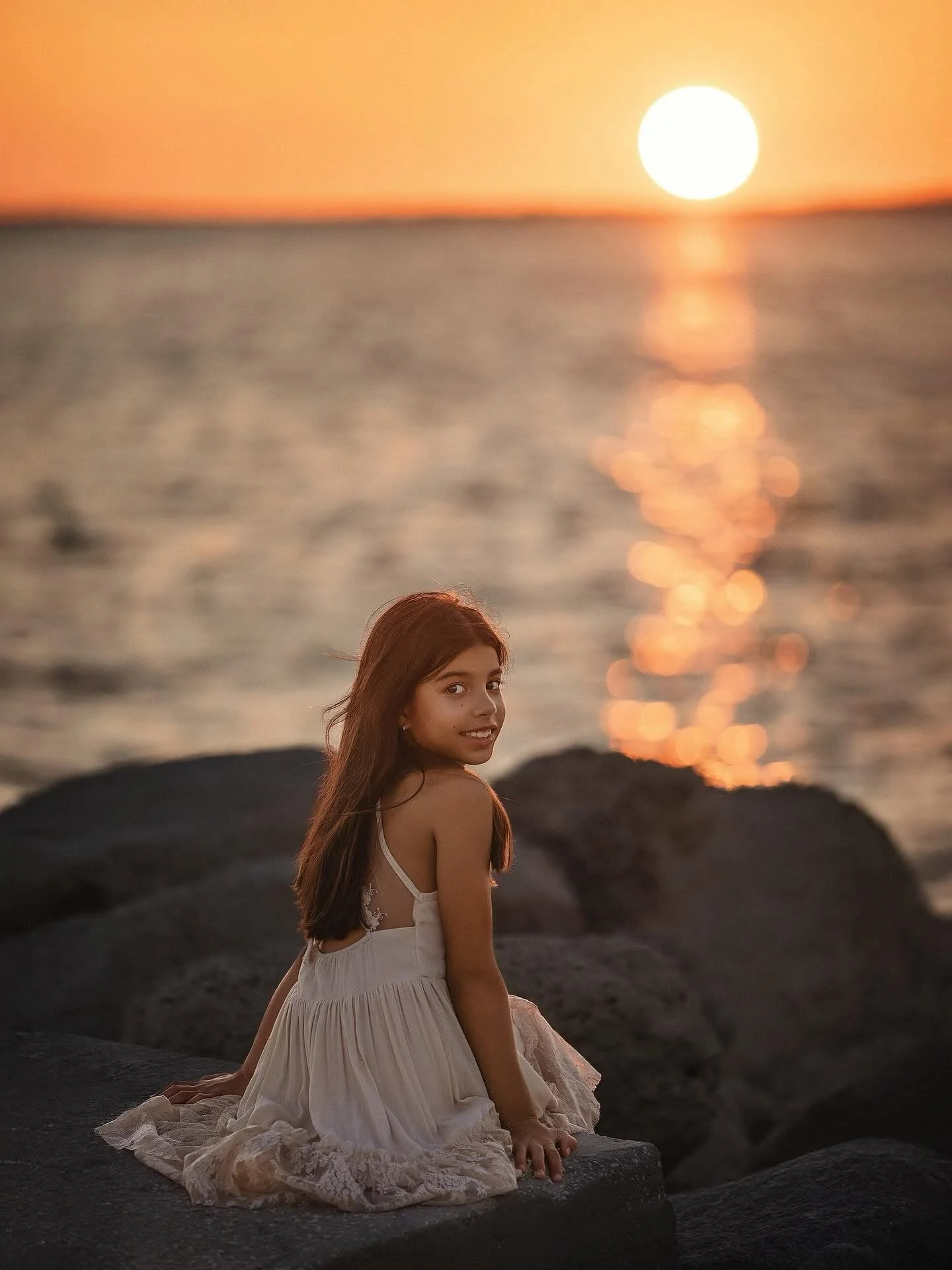 A young woman with long brown hair in a white dress sitting on dark rocks at sunset by the ocean, with the sun reflecting on the water.