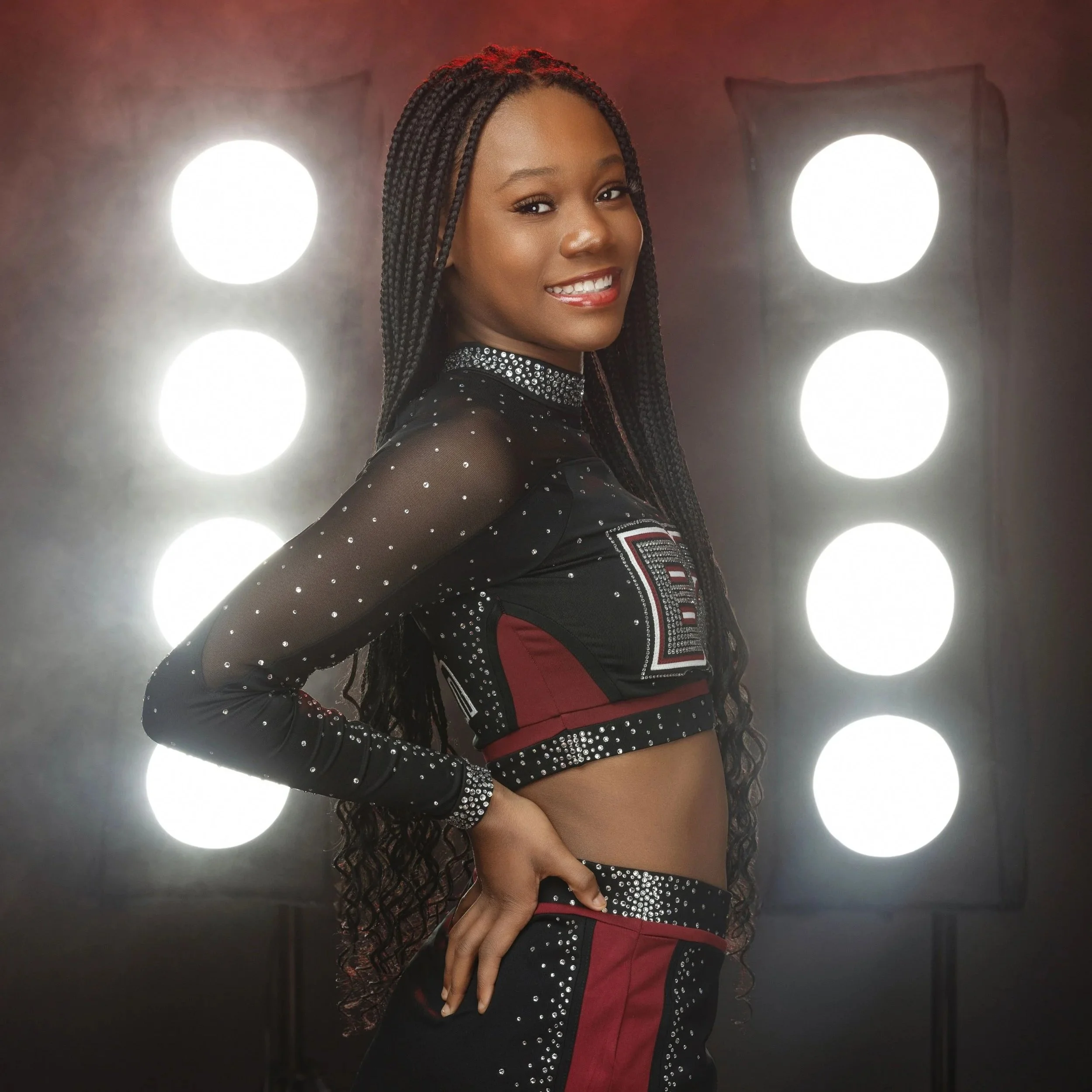 Woman with long braided hair smiling, wearing a black and red sports outfit with rhinestone embellishments, standing in front of circular bright lights.