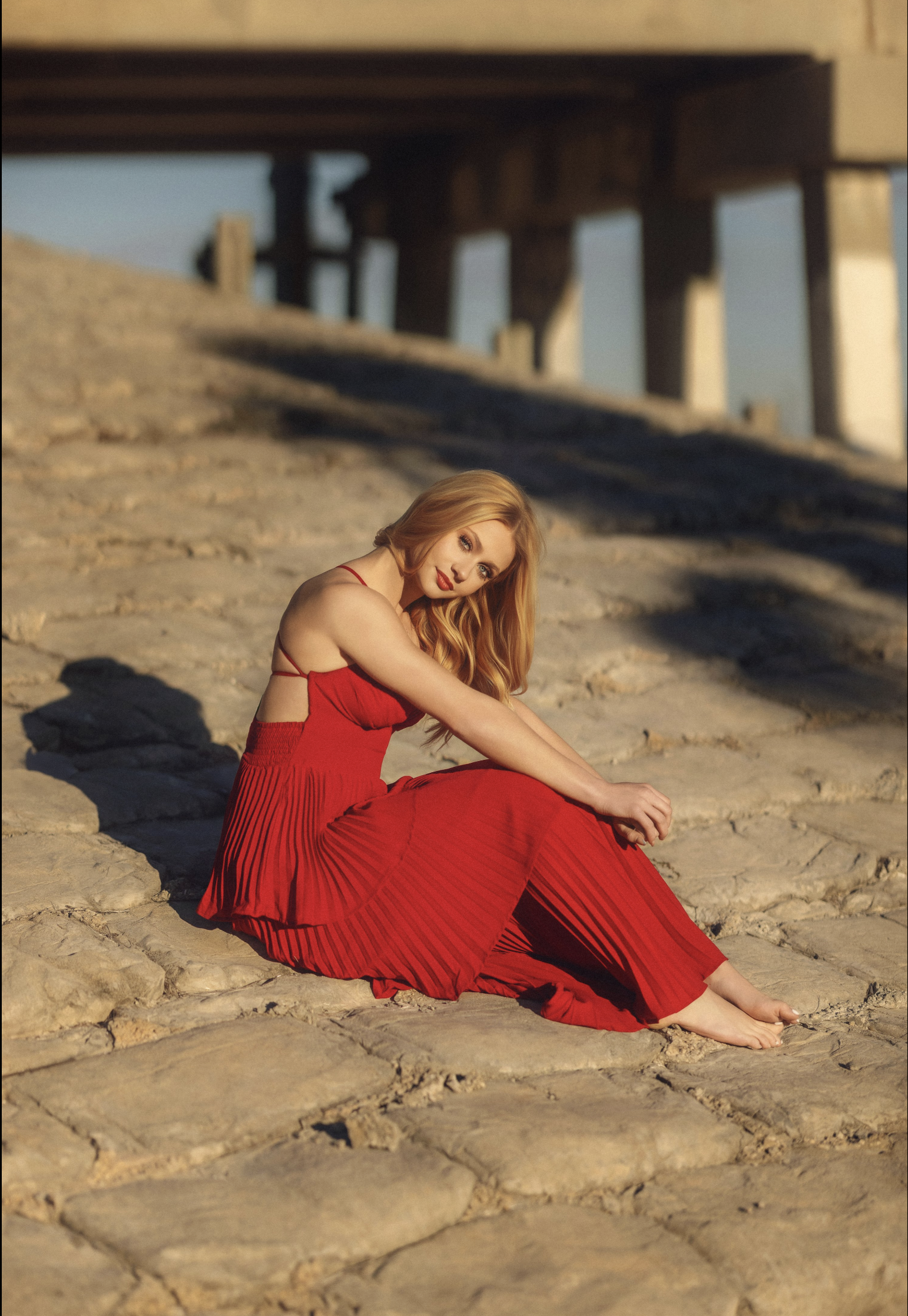 A woman with long red hair sitting on a sandy beach, wearing a red dress, with a bridge in the background.
