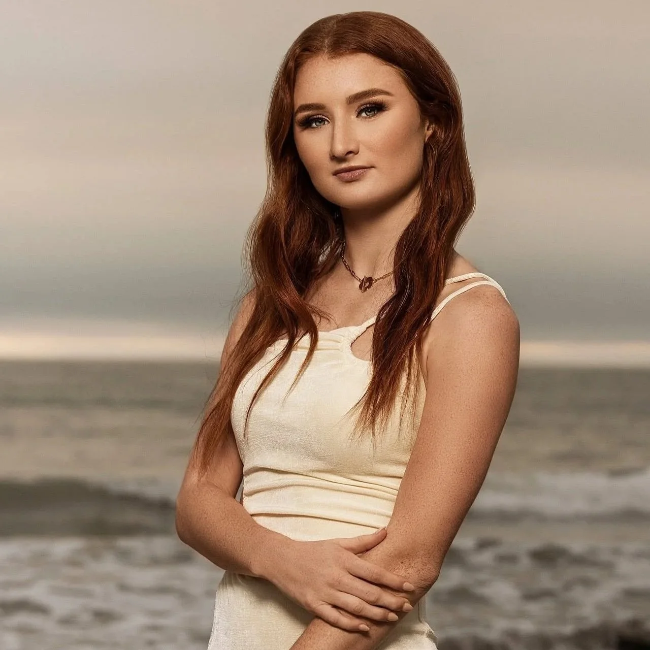A young woman with long red hair and fair skin standing on a beach, wearing a cream-colored sleeveless dress and a small necklace, with a calm ocean and cloudy sky in the background.