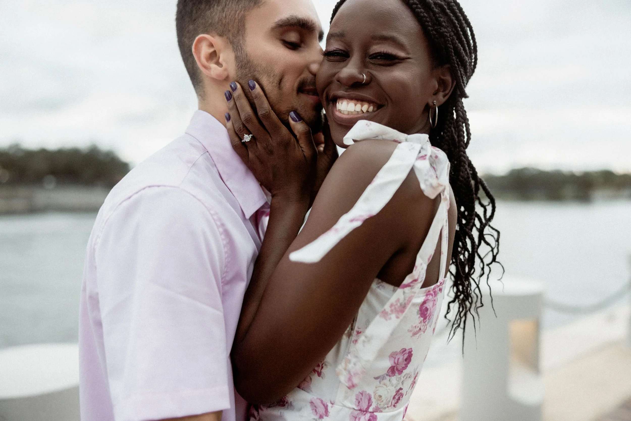 A joyful couple embraces on a beach, with the man whispering into the woman's ear. The woman is smiling widely, showing her teeth, and wearing hoop earrings and a floral dress. The man has a short haircut and a beard, and is wearing a light pink shirt. The background is blurred with water and trees visible.