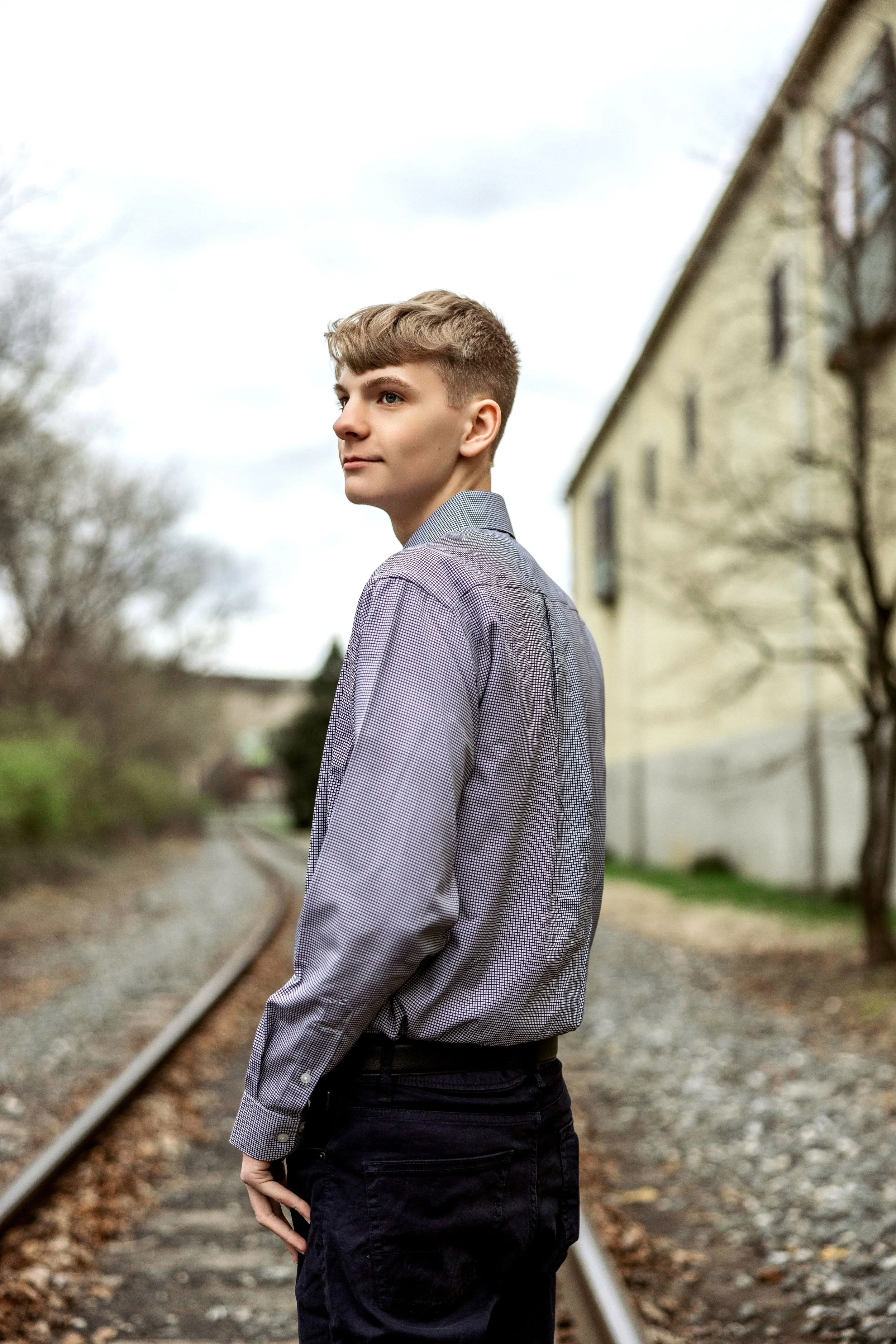 A young man with short light brown hair standing outdoors on train tracks, wearing a light gray checkered shirt and black jeans, looking to the side with a serious expression, alongside a yellowish building and leafless trees.