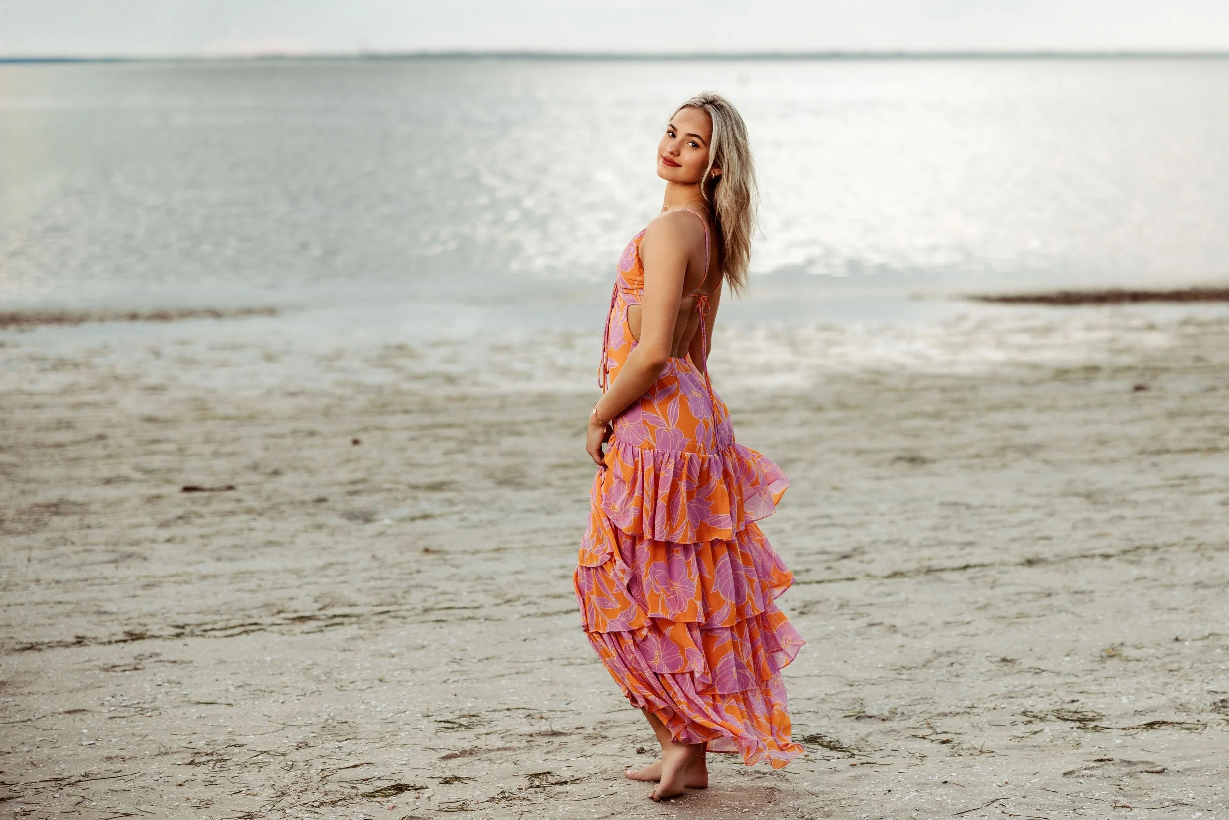 A woman with long blonde hair wearing a colorful floral dress stands barefoot on the sandy beach near the water, smiling gently as she looks into the camera.