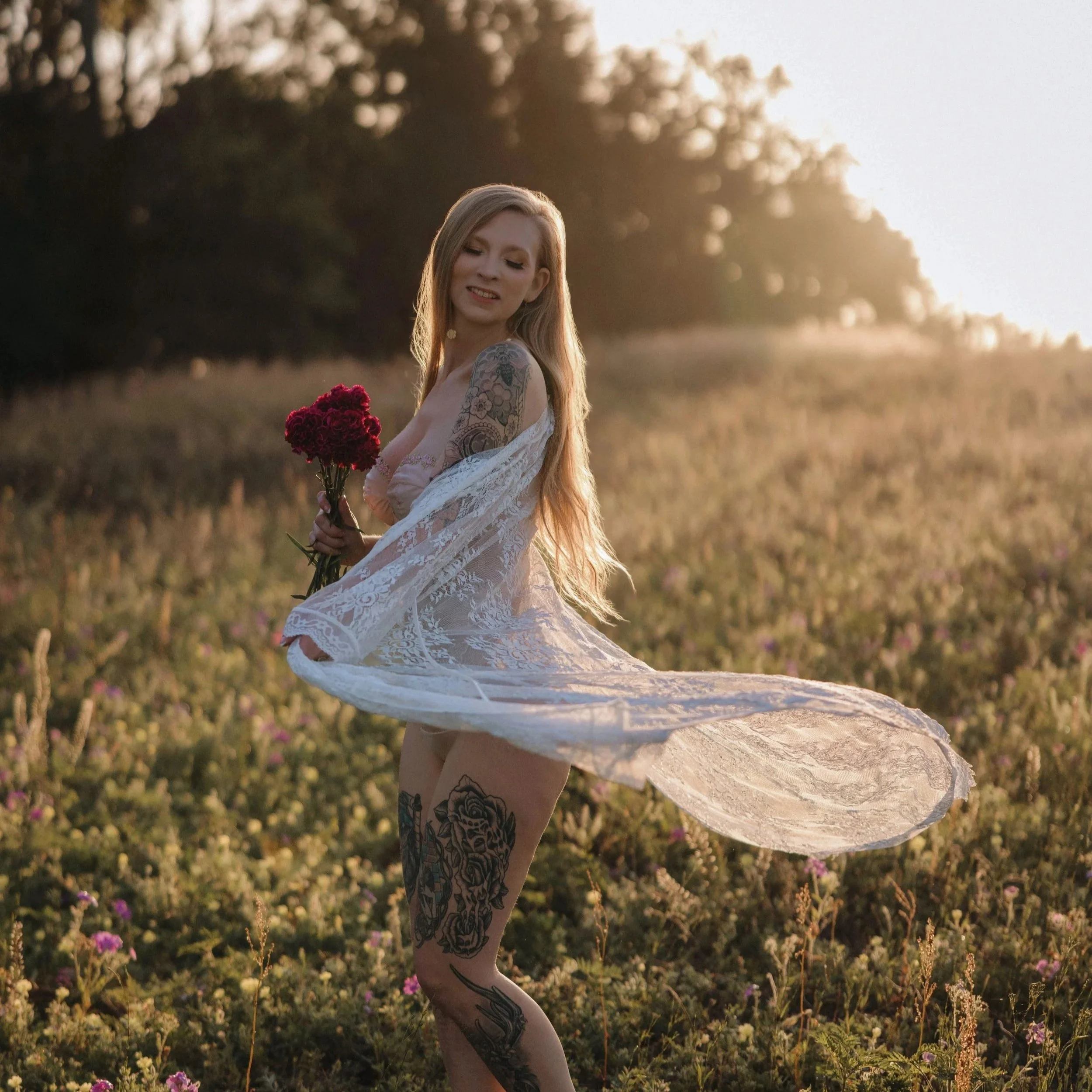 A woman with long blonde hair and tattoos dressed in a white lace dress standing in a field with flowers during sunset, holding a bouquet of red flowers and smiling.