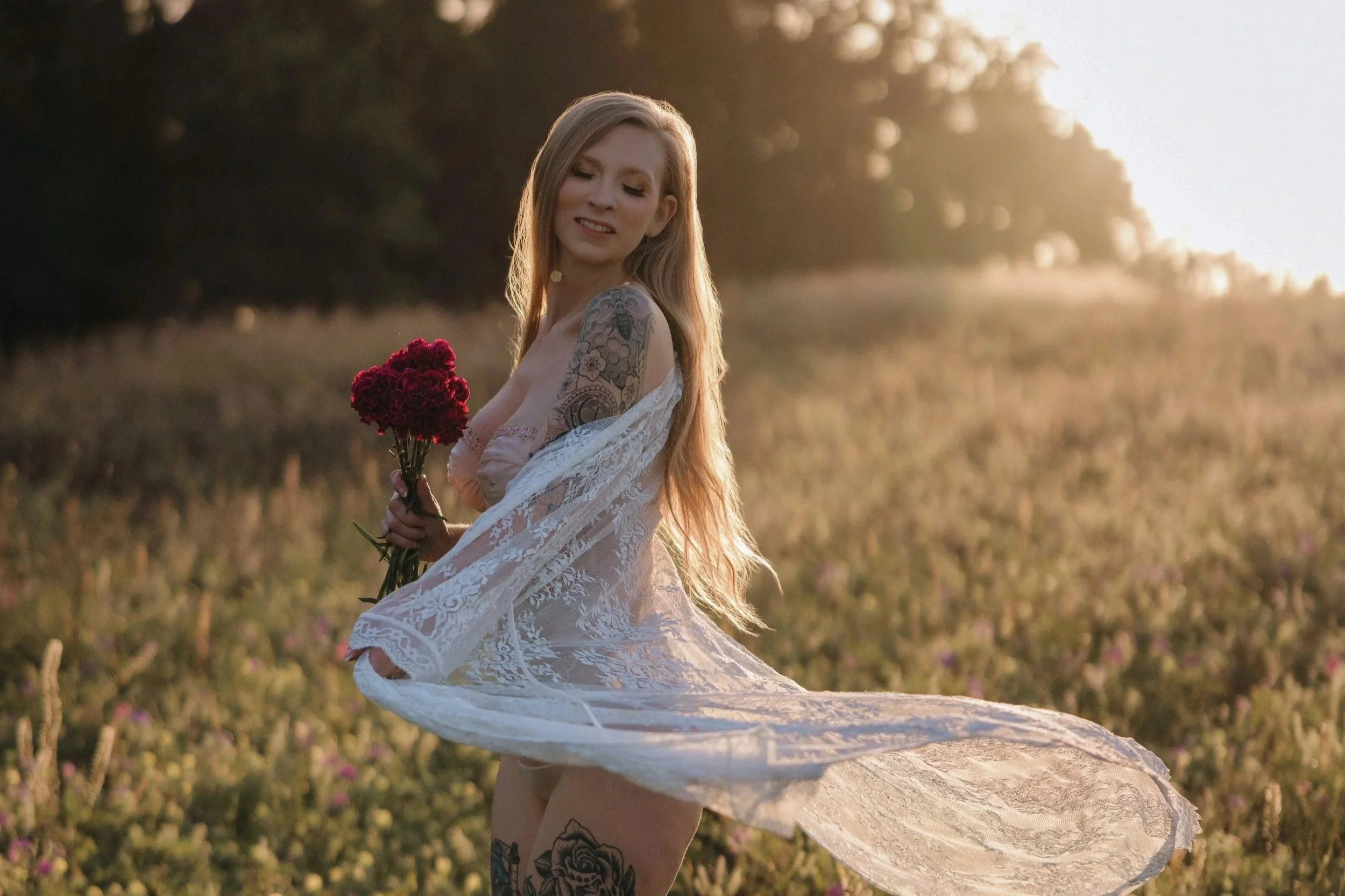 A woman with long blonde hair, tattoos on her arm and thigh, wearing a white lace dress, standing in a field of grass and wildflowers during sunset, holding a bouquet of red flowers.