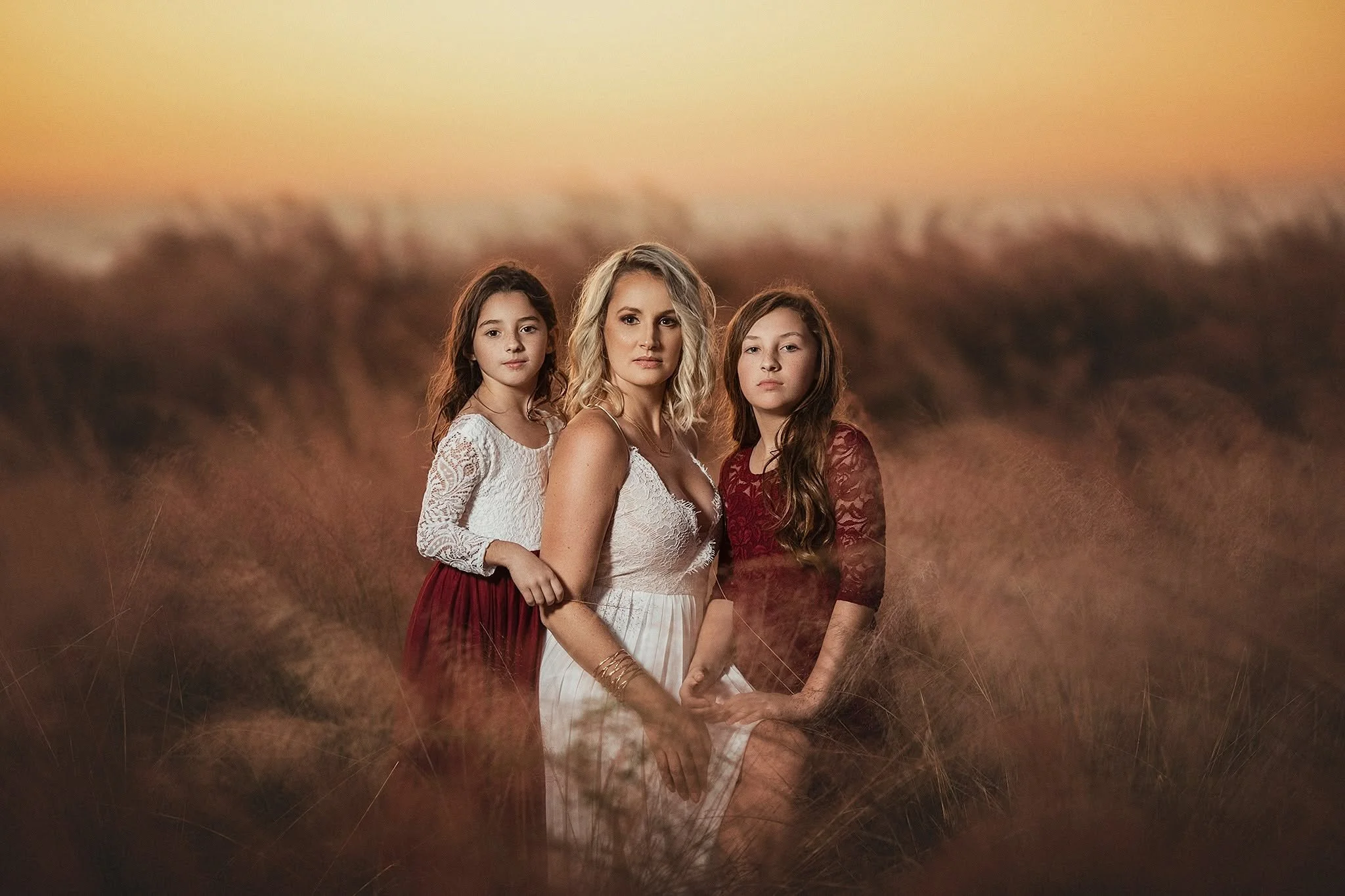 Three women standing in a field during sunset, with the sky glowing orange in the background.