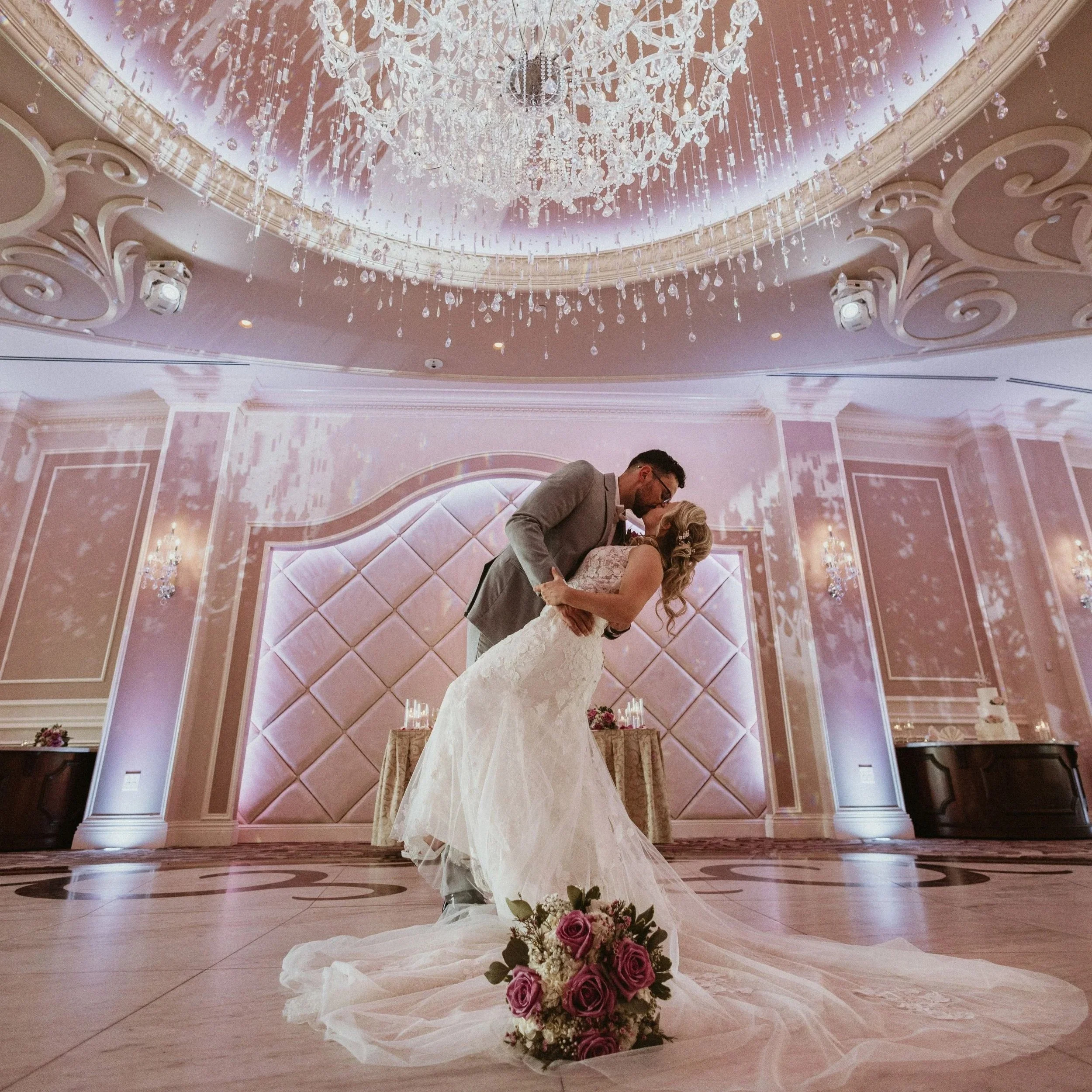 A bride and groom dance in a luxurious, ornately decorated ballroom with a chandelier hanging from a pink ceiling. The bride wears a white wedding gown and the groom wears a gray suit. A bouquet of pink and white roses is placed on the floor at the forefront.