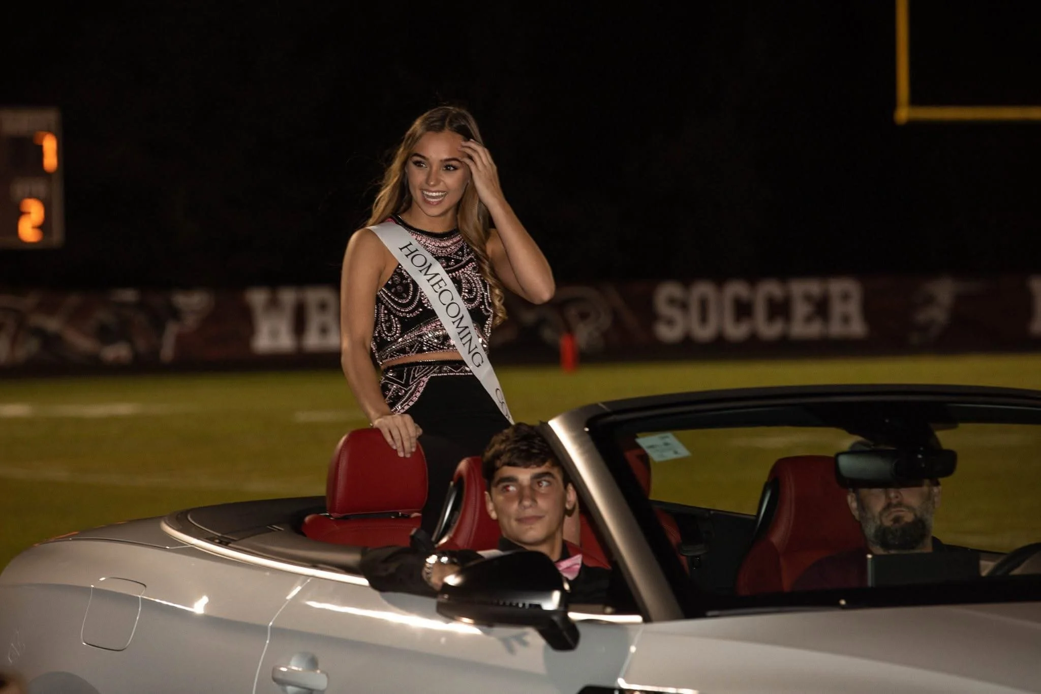 A woman wearing a sash that says "Homecoming" is standing in the back seat of a silver convertible car during a night event. She is smiling and touching her head, surrounded by other people in the car.