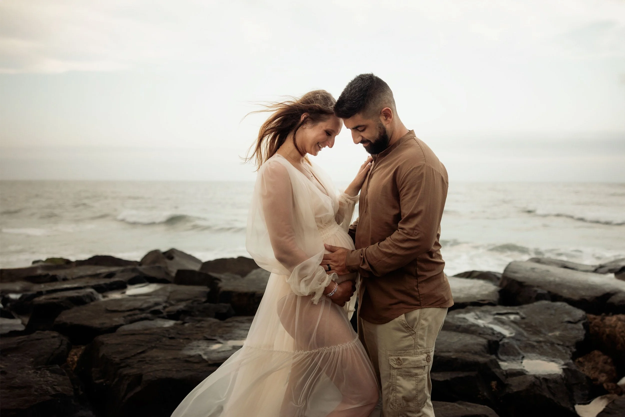 A couple standing close together on a rocky beach, smiling and touching foreheads with their eyes closed under a cloudy sky.