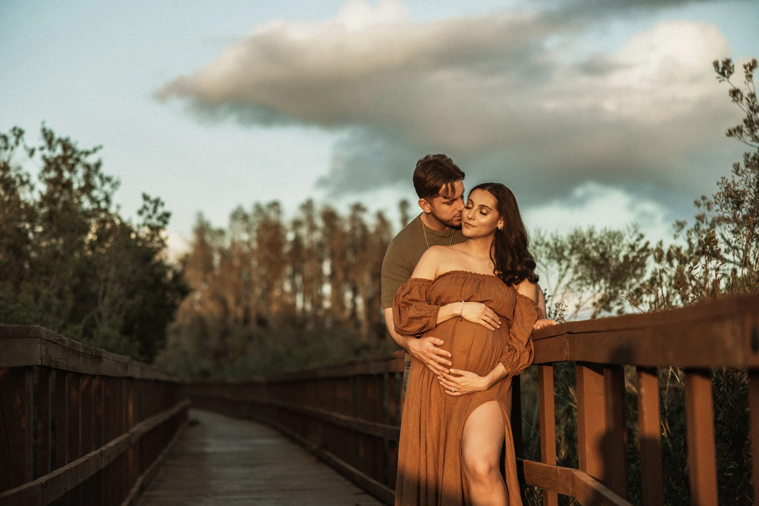 A couple standing on a wooden bridge, with the man embracing the woman from behind, both with closed eyes, during sunset or dusk, surrounded by trees and a cloudy sky in the background.