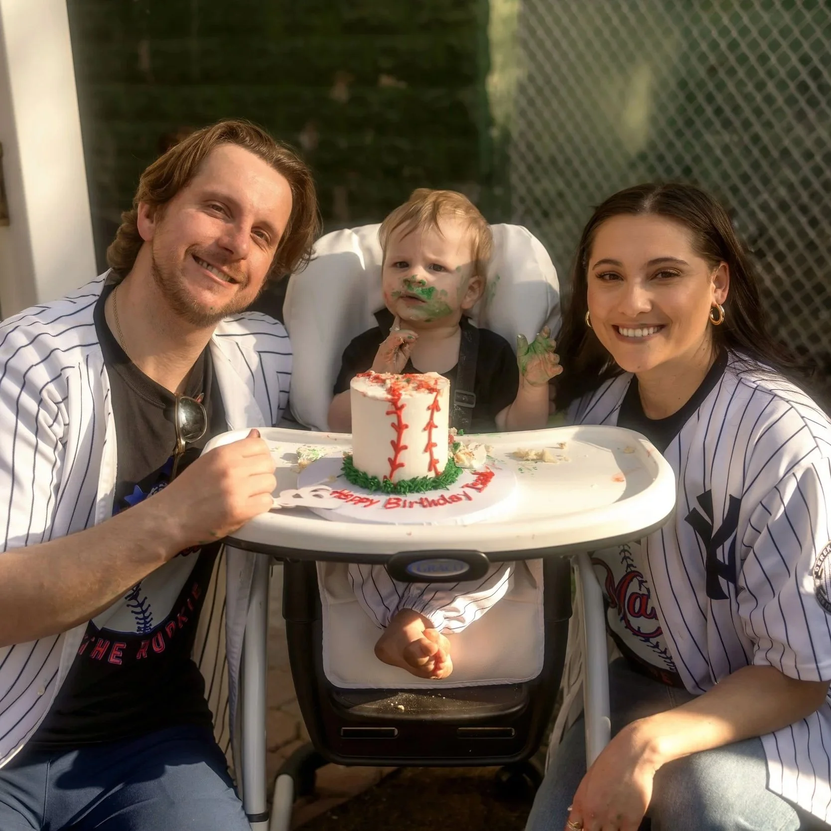 Three people celebrating a birthday outdoors: a man, a young child in a high chair, and a woman, all smiling. The child has green cake on face and hands, with a small baseball-themed birthday cake in front of the child on a tray.