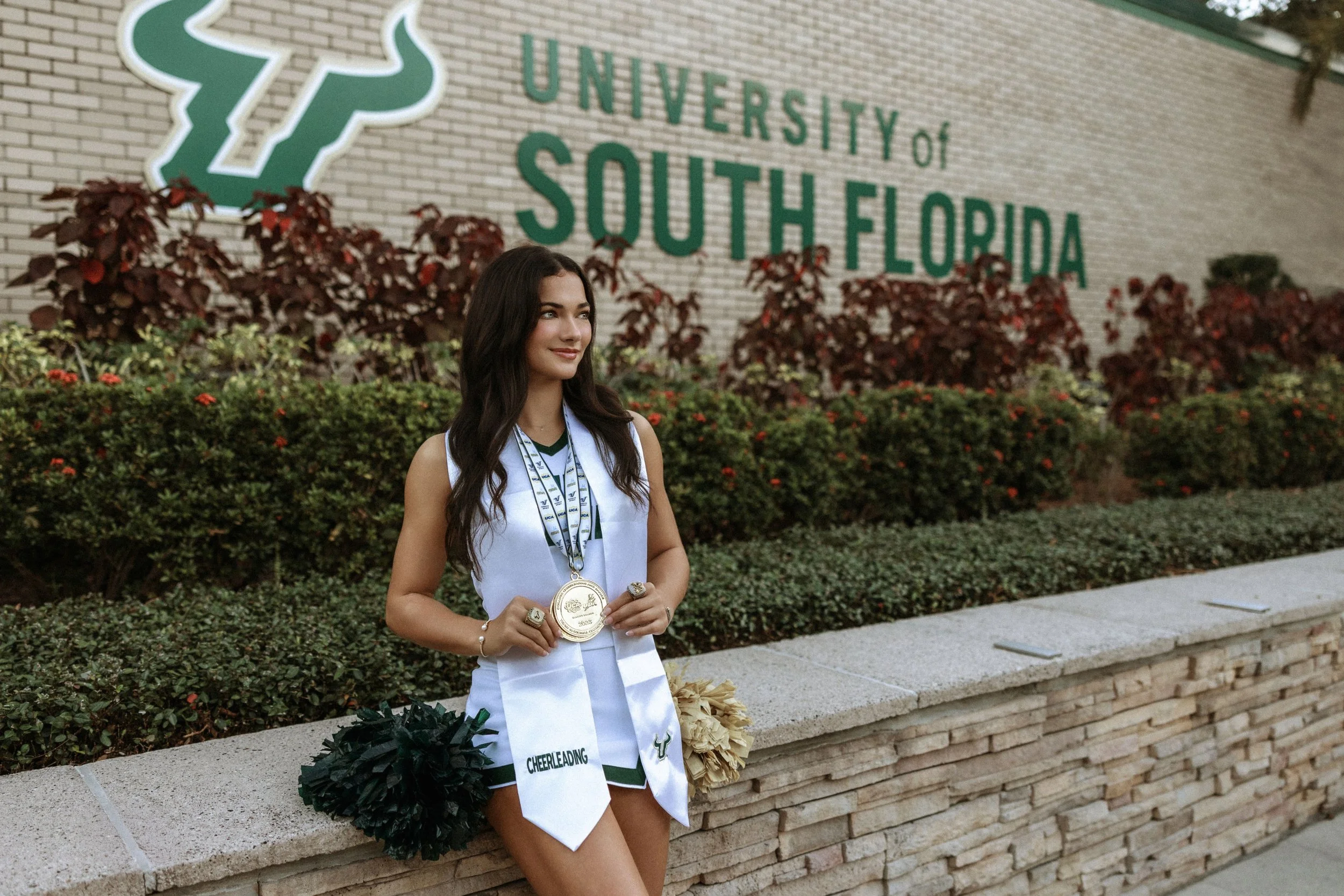 A young woman with long dark hair stands outdoors in front of a campus sign for the University of South Florida, holding medals and wearing a cheerleading uniform.