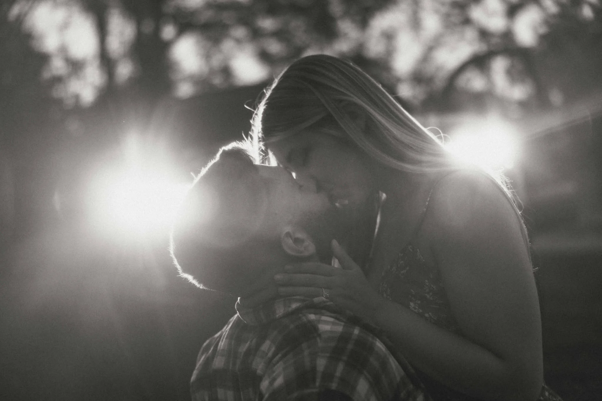 A black and white photo of two people kissing, with the viewpoint from someone standing behind them. One person's hand is visible on the other's back. The background is blurry with lights.