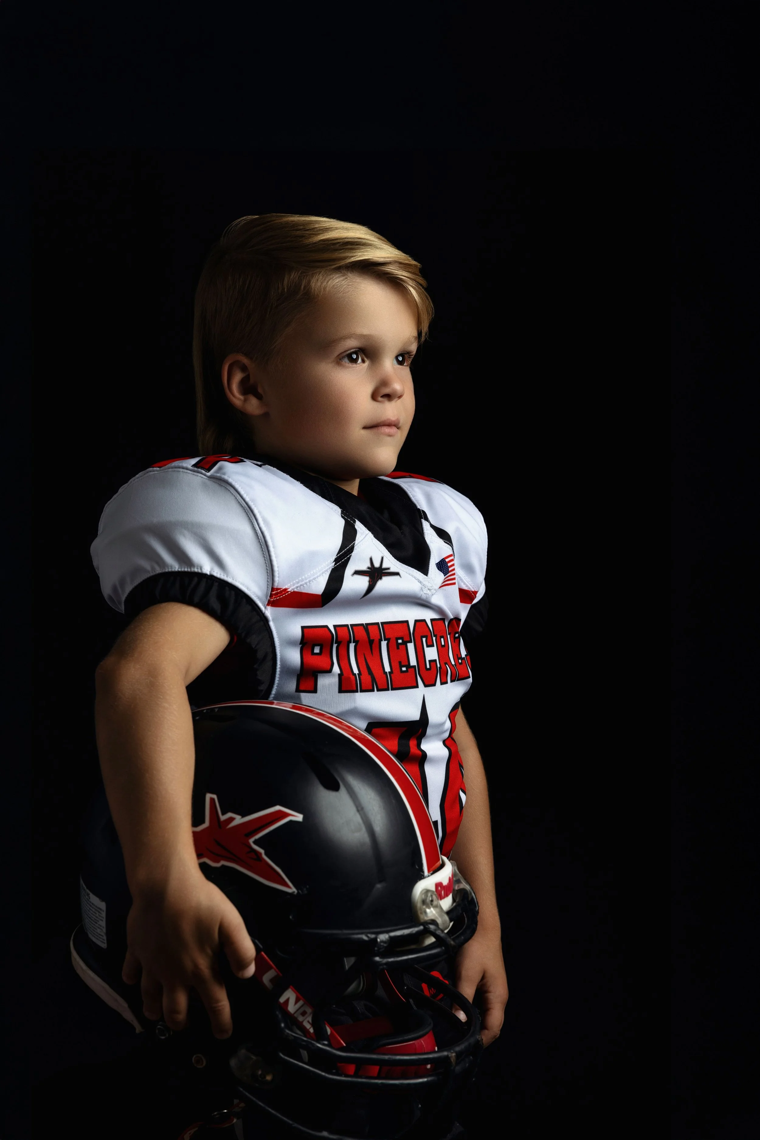 Young boy wearing a football jersey and holding a football helmet, standing against a black background.
