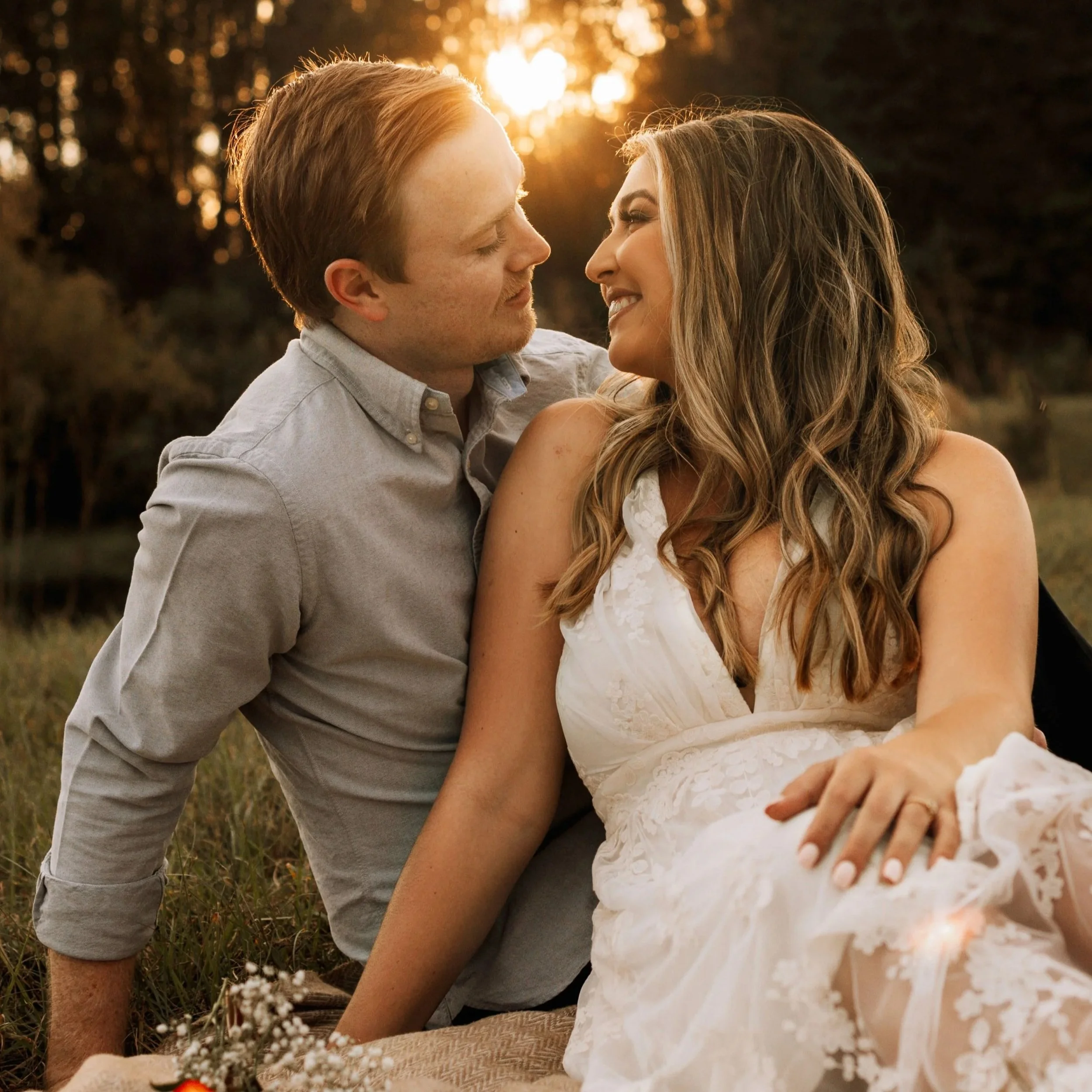A romantic scene of a couple with light skin, sitting close together outdoors at sunset, smiling and looking into each other's eyes. The man has short red hair and is wearing a light-colored shirt, while the woman has long wavy hair and is wearing a white floral dress. The warm sunset light creates a golden glow around them.