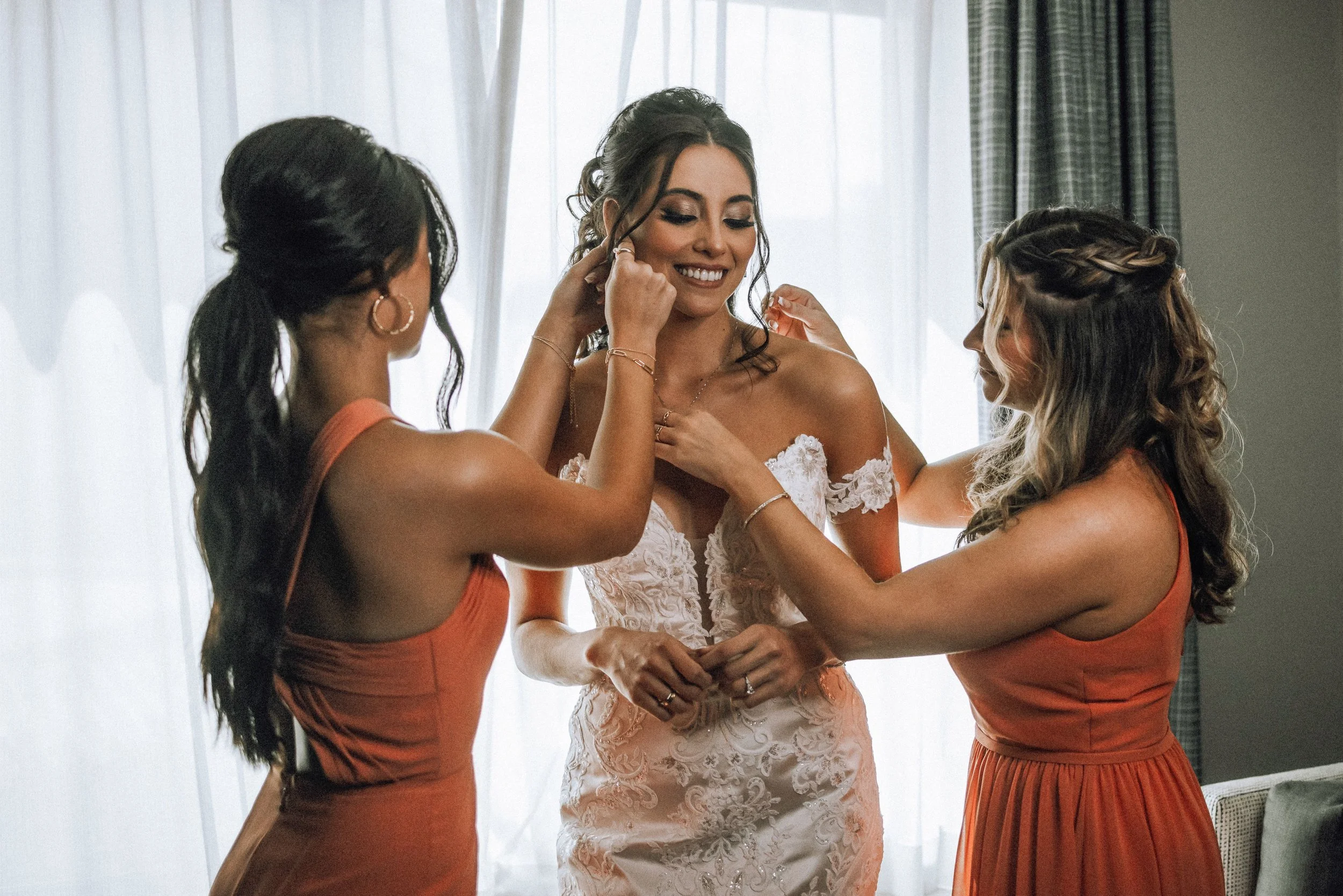 A bride getting ready with three women, possibly bridesmaids, helping her with her wedding dress, smiling in a well-lit room with curtains.