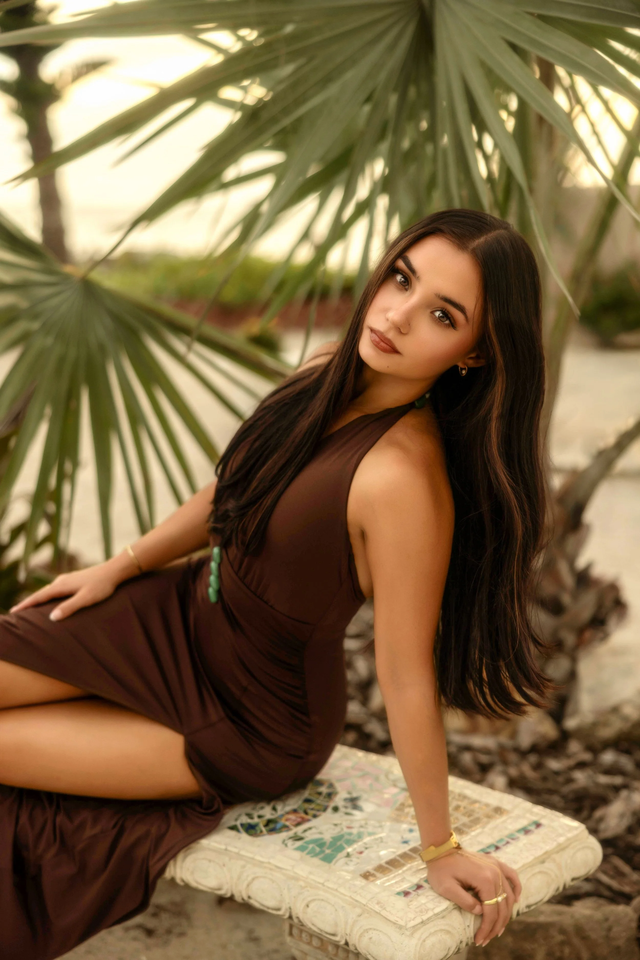 A woman with long dark hair wearing a brown dress, sitting on a bench with a colorful mosaic top and a white base, outdoors near green palm leaves, during what appears to be sunset.