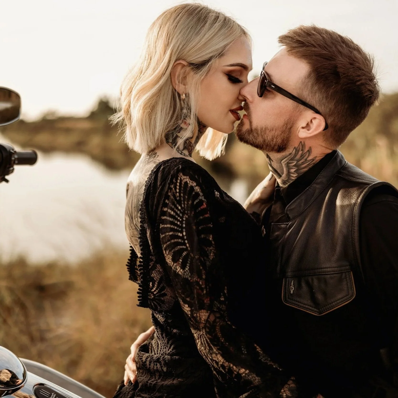 A man and woman are close together outdoors, about to kiss, with a motorcycle handlebar visible in the foreground. The woman has platinum blonde hair, wears makeup, and a black lace top. The man has short brown hair, sunglasses, a beard, tattoos on his neck, and wears a black leather vest.