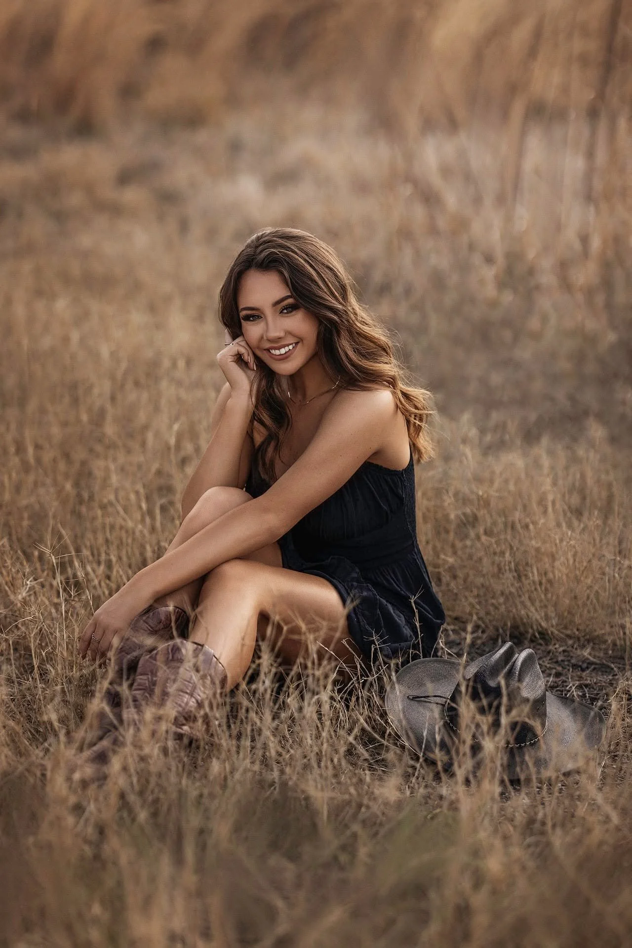 Young woman sitting in a field of tall grass, smiling and looking at the camera, wearing a black dress, cowboy boots, and a bracelet, with her hand resting on her knee.