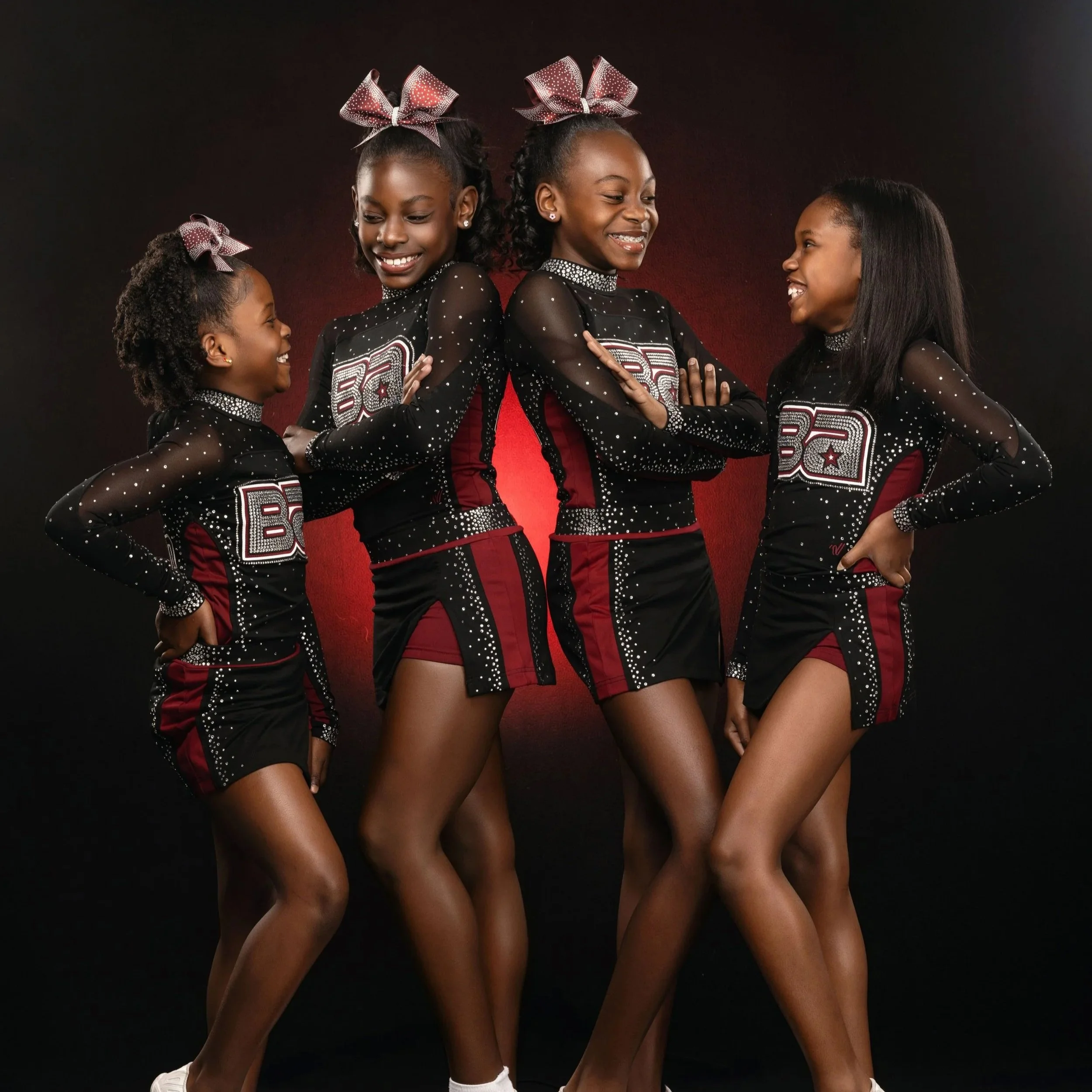 Group of four young African American girls in matching black and red cheerleading uniforms with rhinestones, standing close together, smiling and laughing against a dark background, with their arms around each other.