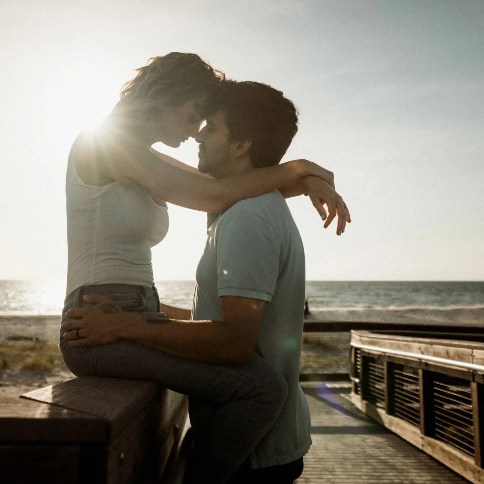 A couple embracing at sunset on a beach, with the woman sitting on a wooden bench and the man standing beneath her, with the ocean and sky in the background.