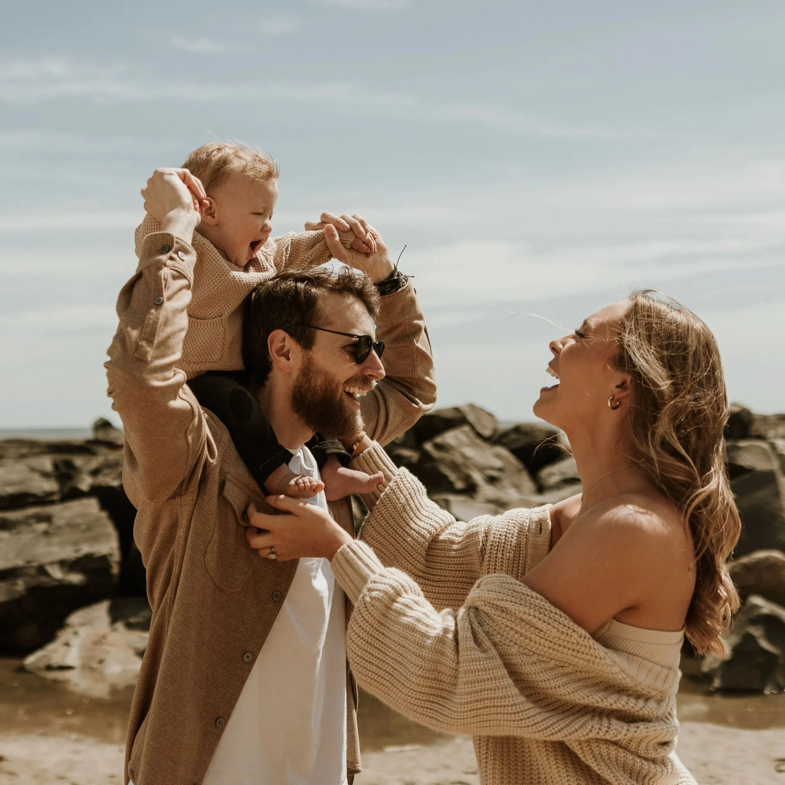 A man and a woman are close together, smiling and looking at each other, while a child sits on the man's shoulders and yawns or sings, at the beach with rocks and cloudy sky in the background.