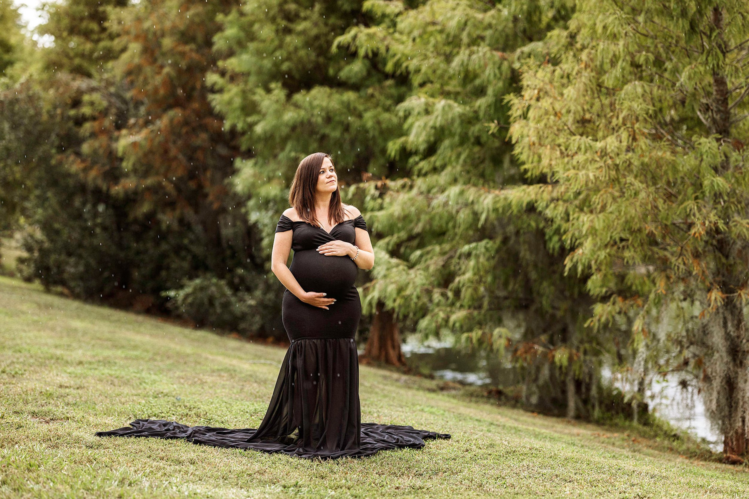 A pregnant woman in a black dress stands outdoors on grass near trees, holding her belly and looking contemplative. A black fabric is spread on the ground around her.