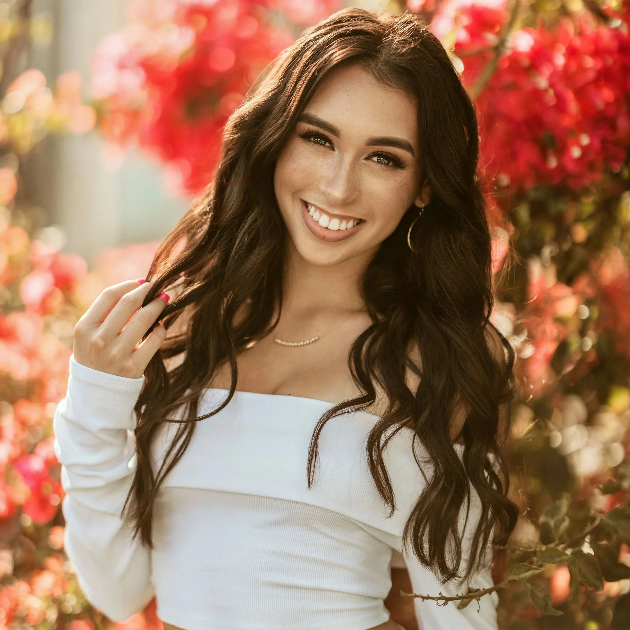 A woman with long dark hair, smiling, wearing a white long-sleeve top, standing outdoors with red autumn leaves in the background.