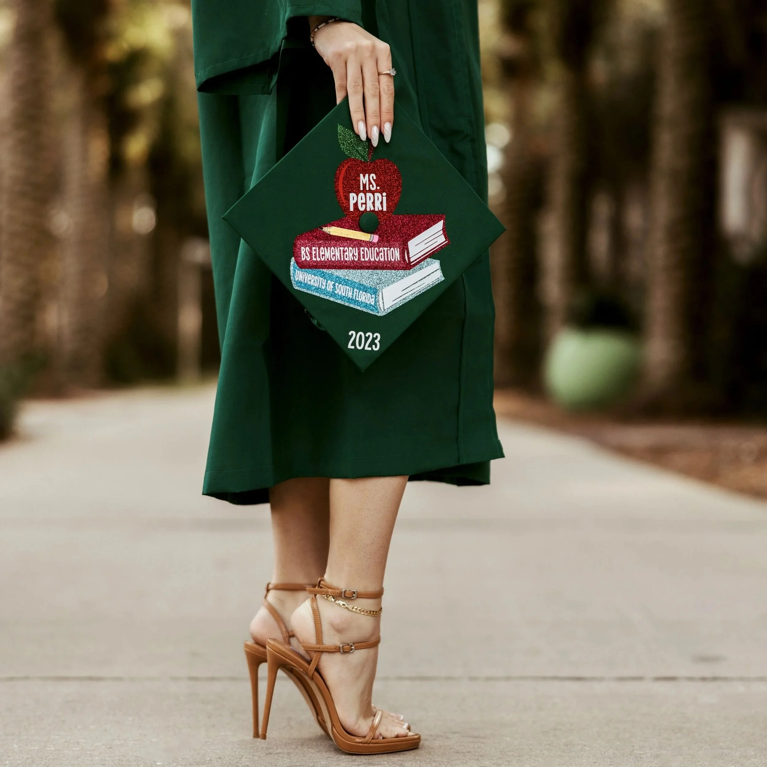 Close-up of a woman’s lower body dressed in a green school uniform skirt, wearing tan high-heeled sandals and an ankle bracelet, standing on a sidewalk.