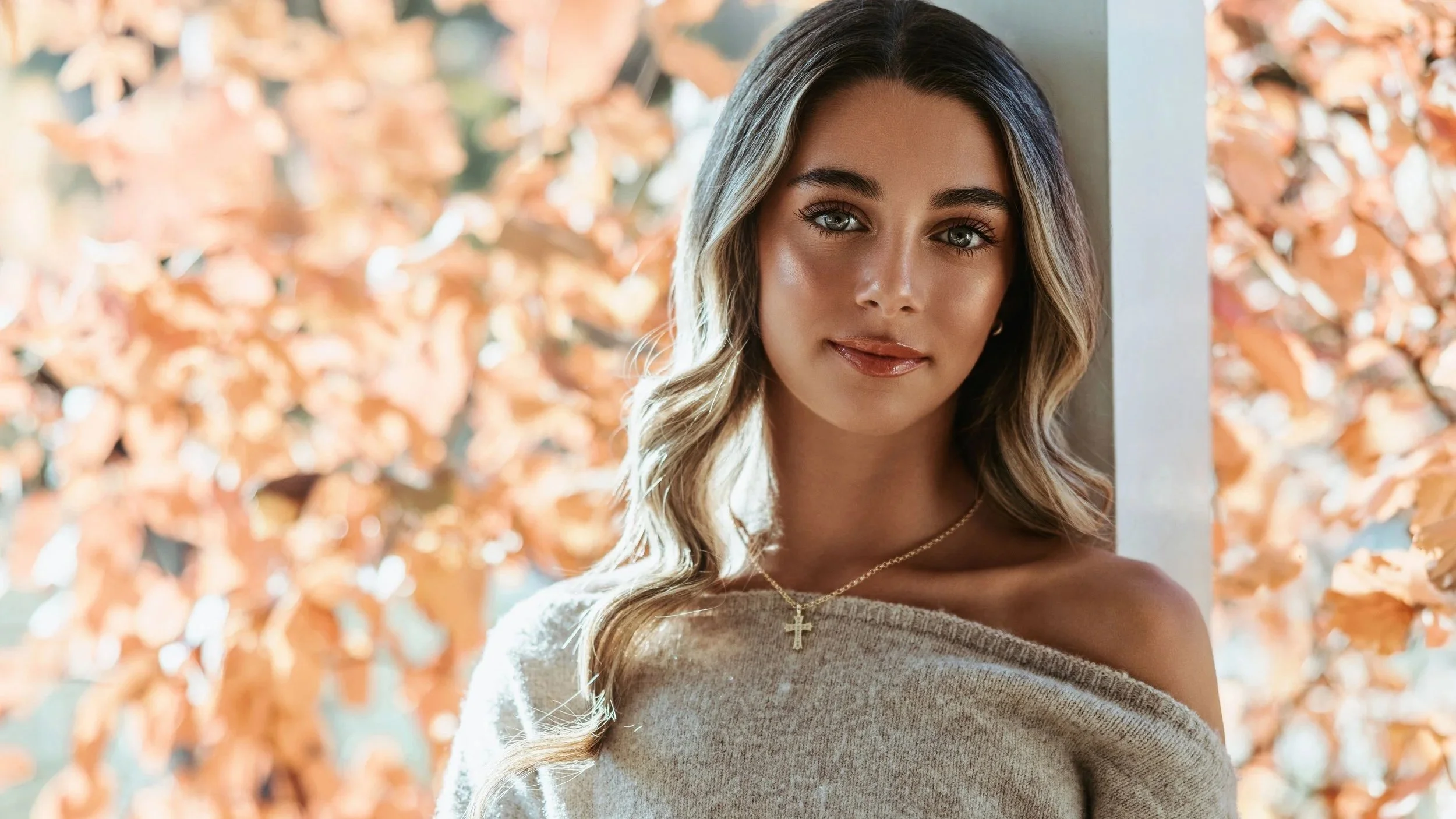 A young woman with long wavy hair, light makeup, and a gold cross necklace, standing in front of orange autumn leaves, looking at the camera with a slight smile.