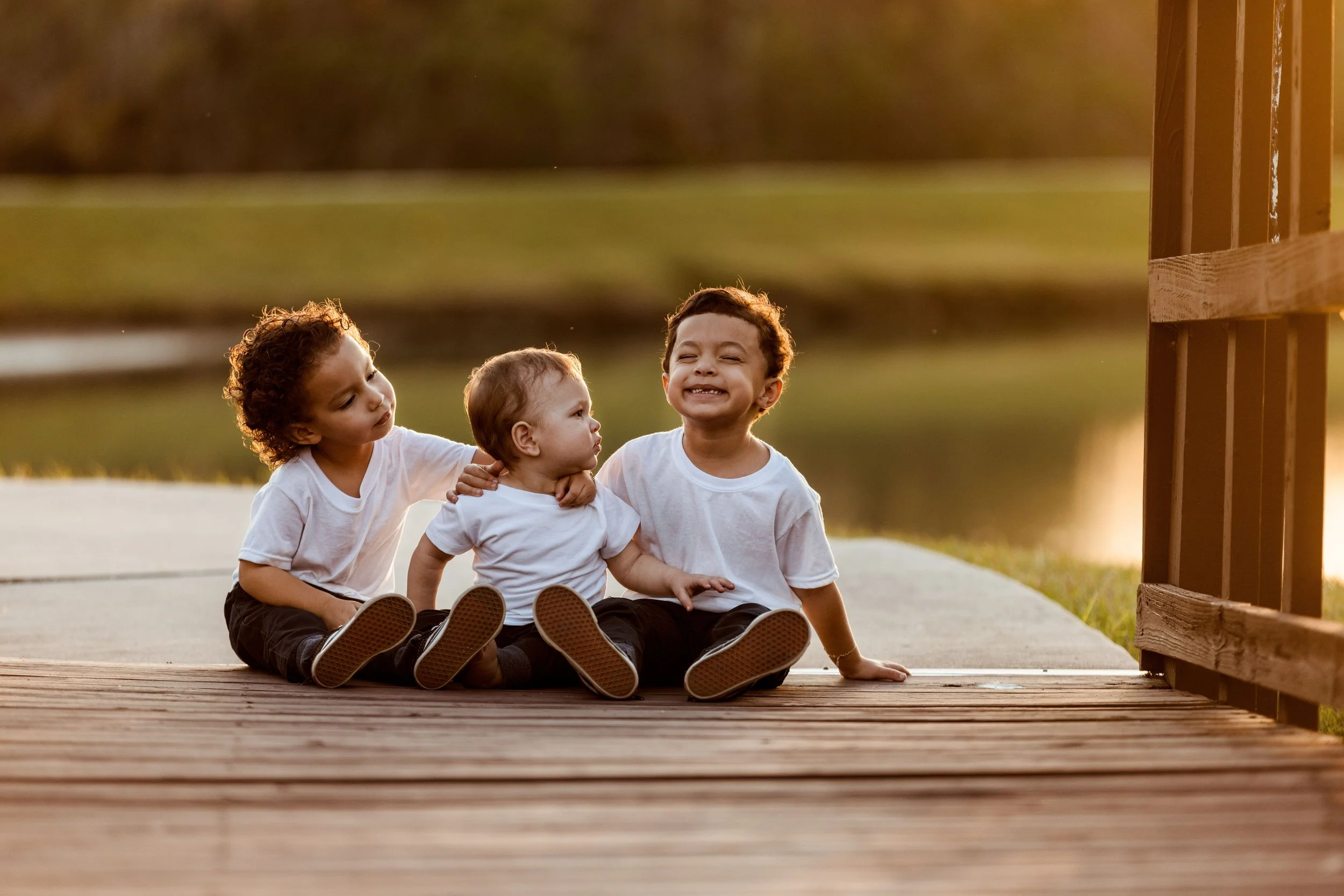 Three young children sitting on a wooden deck outside during sunset, smiling and interacting with each other. One girl leans in to talk to a younger child, while a boy smiles broadly. They are dressed in casual white shirts and black pants, with a grassy field and trees in the background.