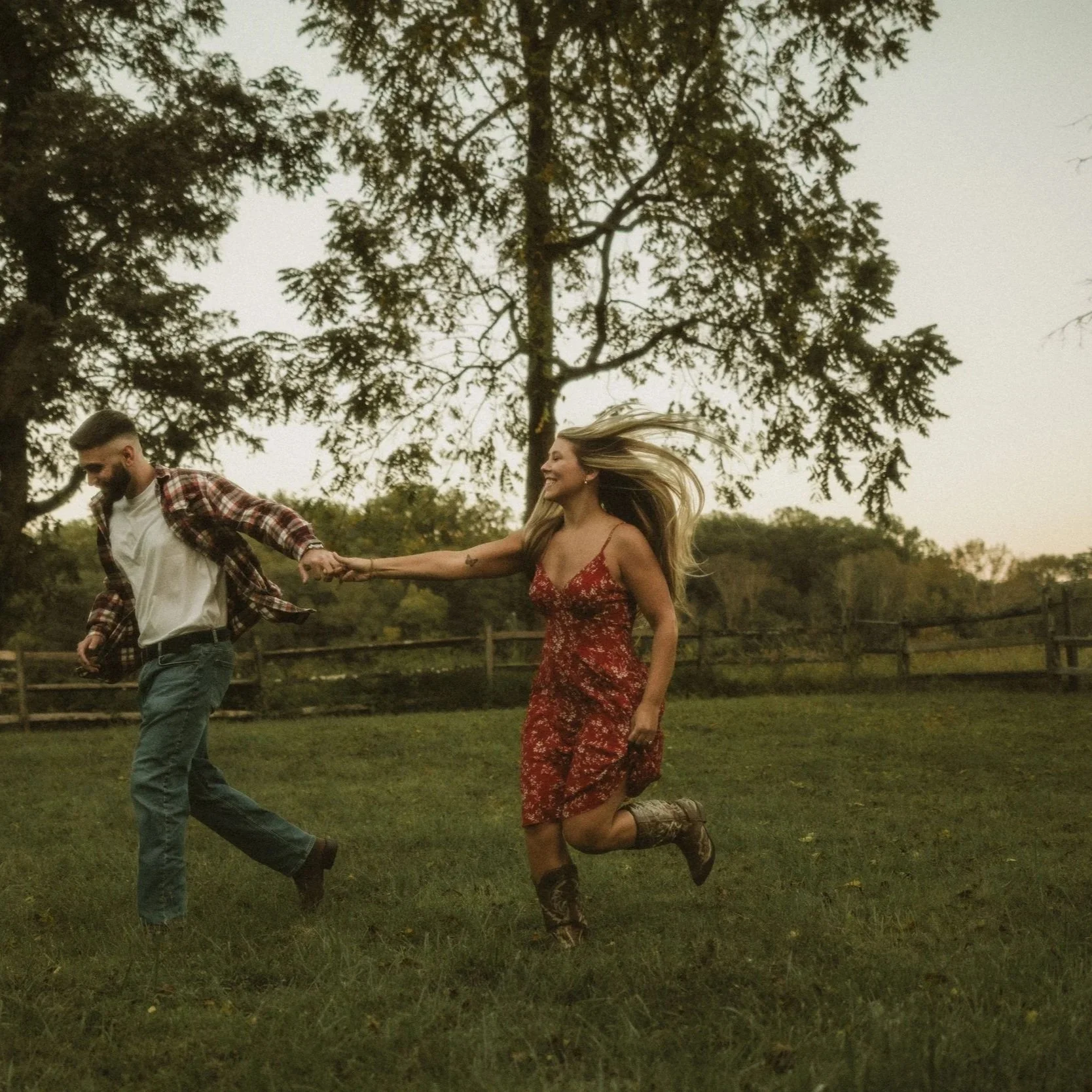 A man and woman holding hands and running through a grassy field, with trees in the background, during what appears to be late afternoon or sunset.