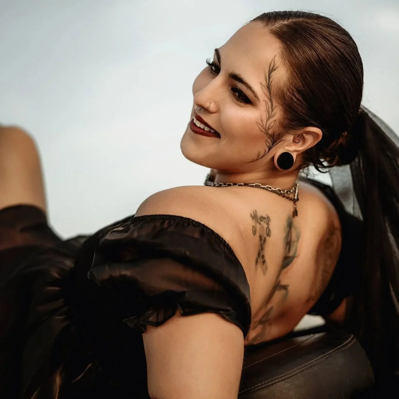 A young woman with tattoos smiling while looking over her shoulder, wearing black earrings, a chain necklace, and a sheer black top, with a plain white background.