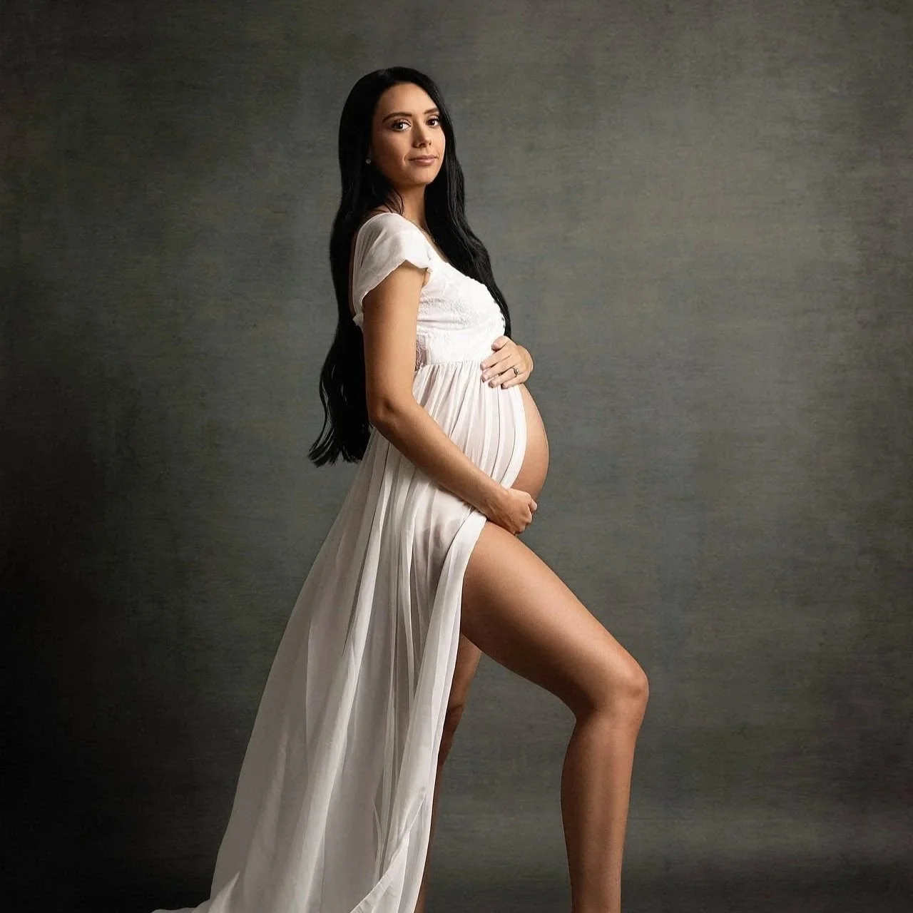 A pregnant woman with long dark hair standing sideways, wearing a white dress, gently holding her belly with one hand and resting her other hand on her thigh against a plain gray background.