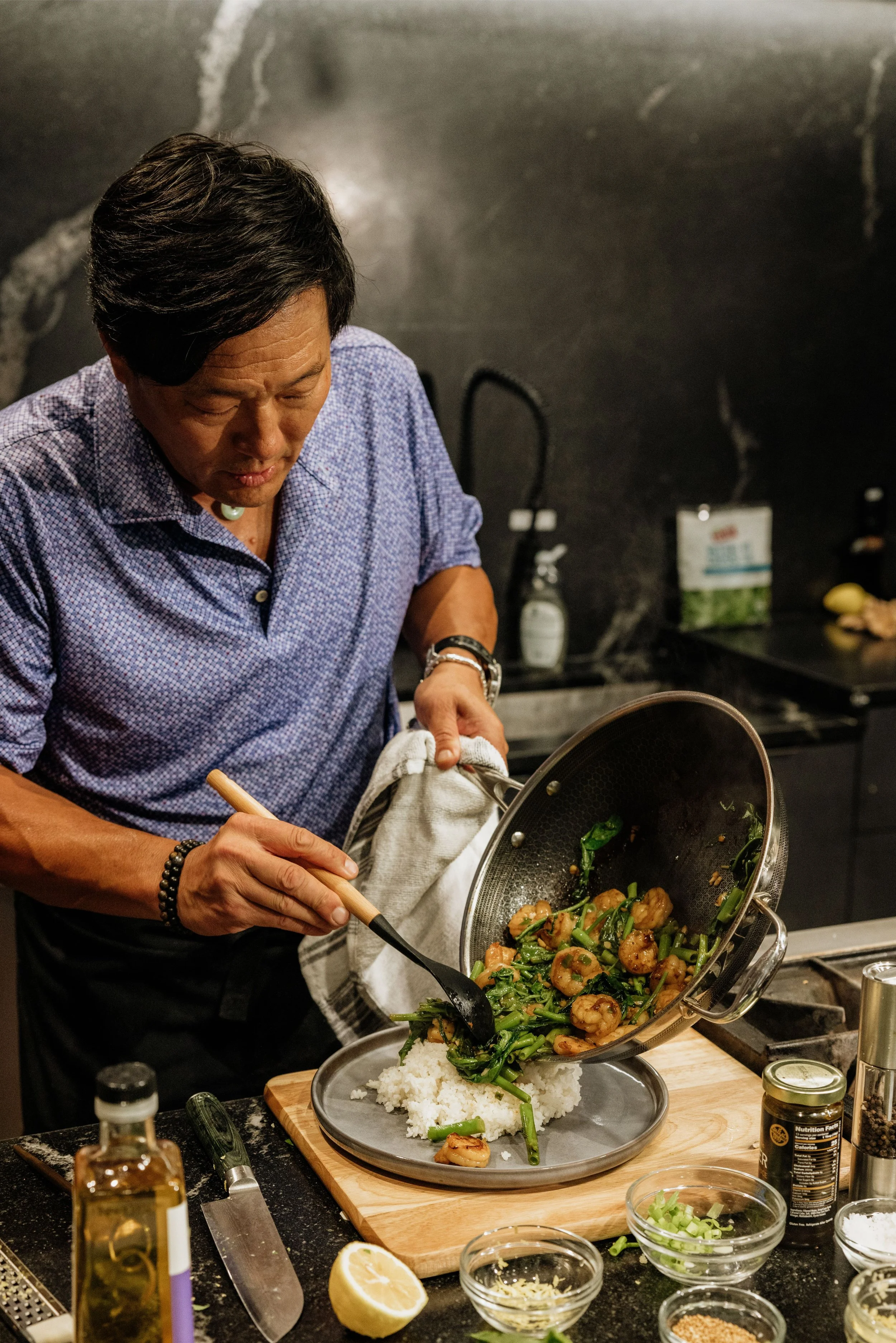 A man cooking in a kitchen, pouring sautéed vegetables and shrimp over rice on a plate.