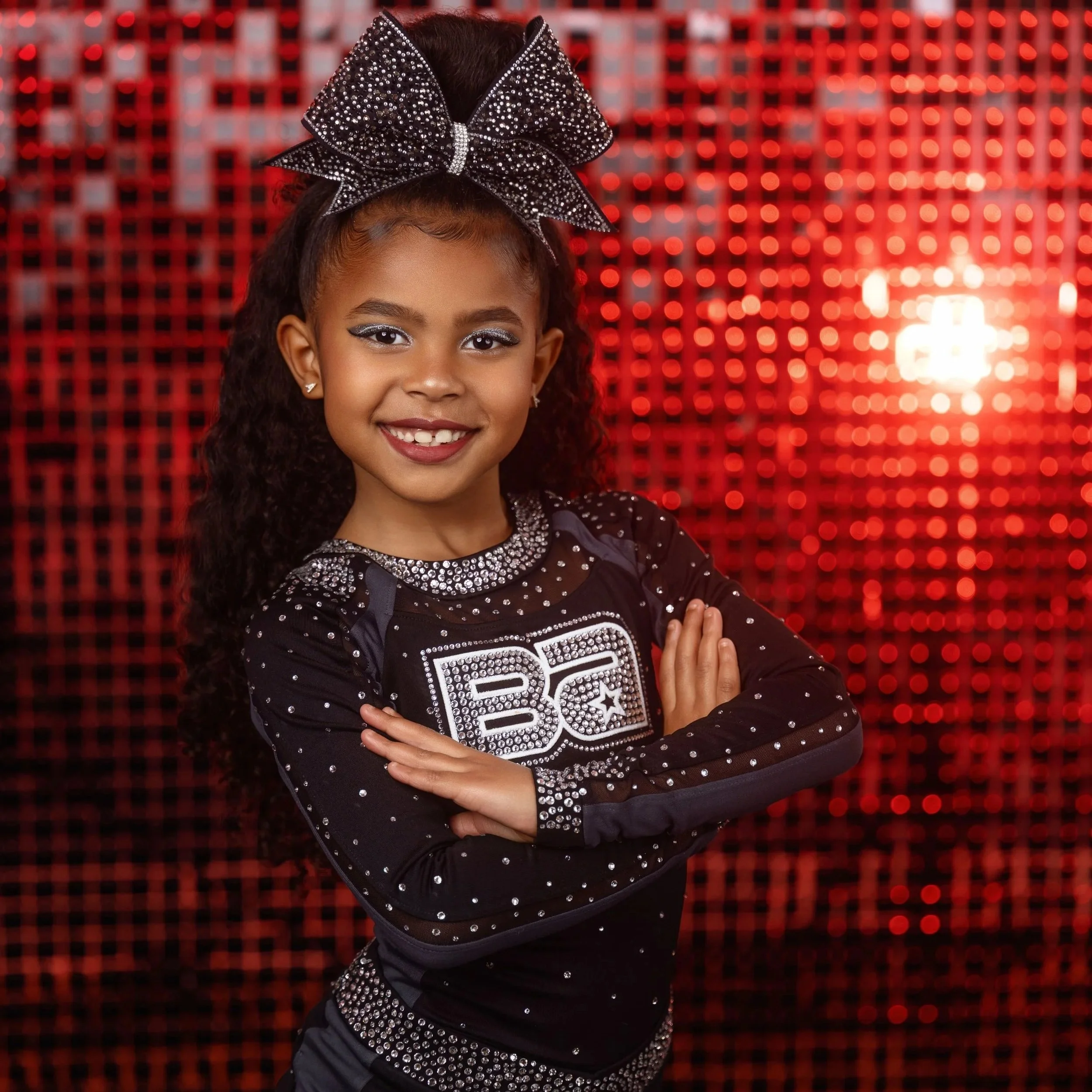 Close-up of a young girl smiling, wearing a black outfit decorated with rhinestones, a large black rhinestone bow on her head, and a red bokeh background.