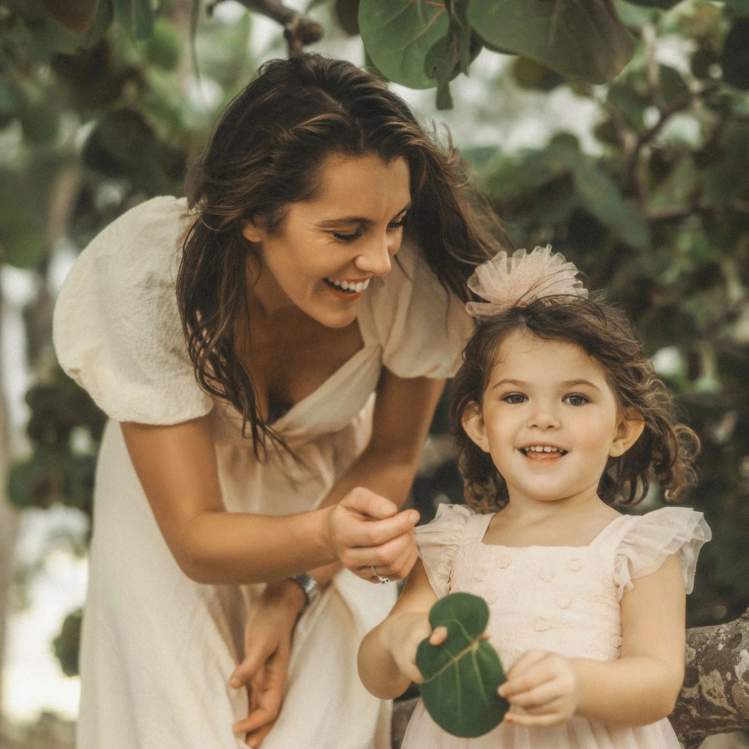 A woman with wavy brown hair smiling and leaning over a young girl with curly brown hair, both wearing light-colored dresses, outdoors surrounded by green leaves.
