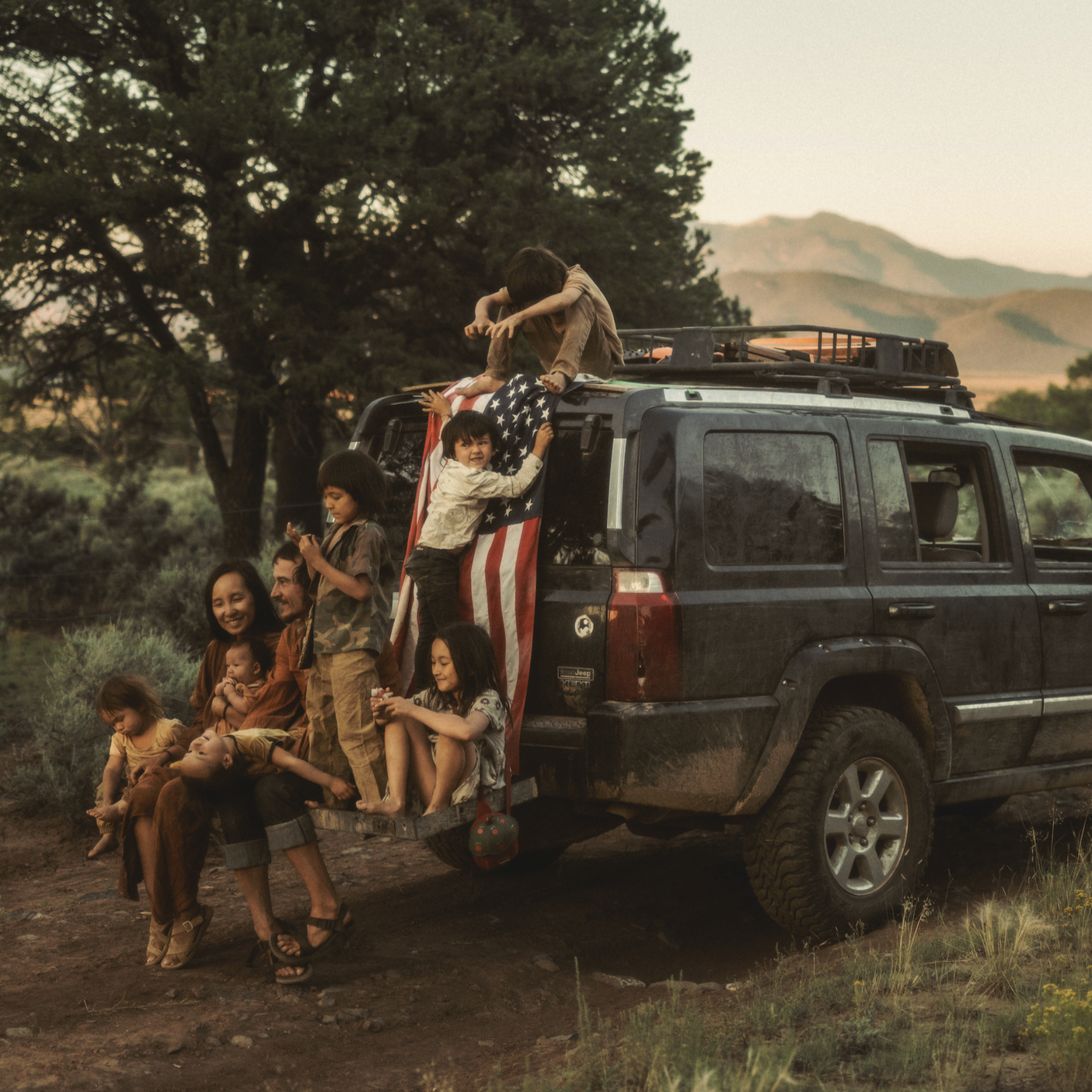 A group of children and a woman sit on the back of a black SUV outdoors during sunset, with a landscape of trees and mountains in the background.
