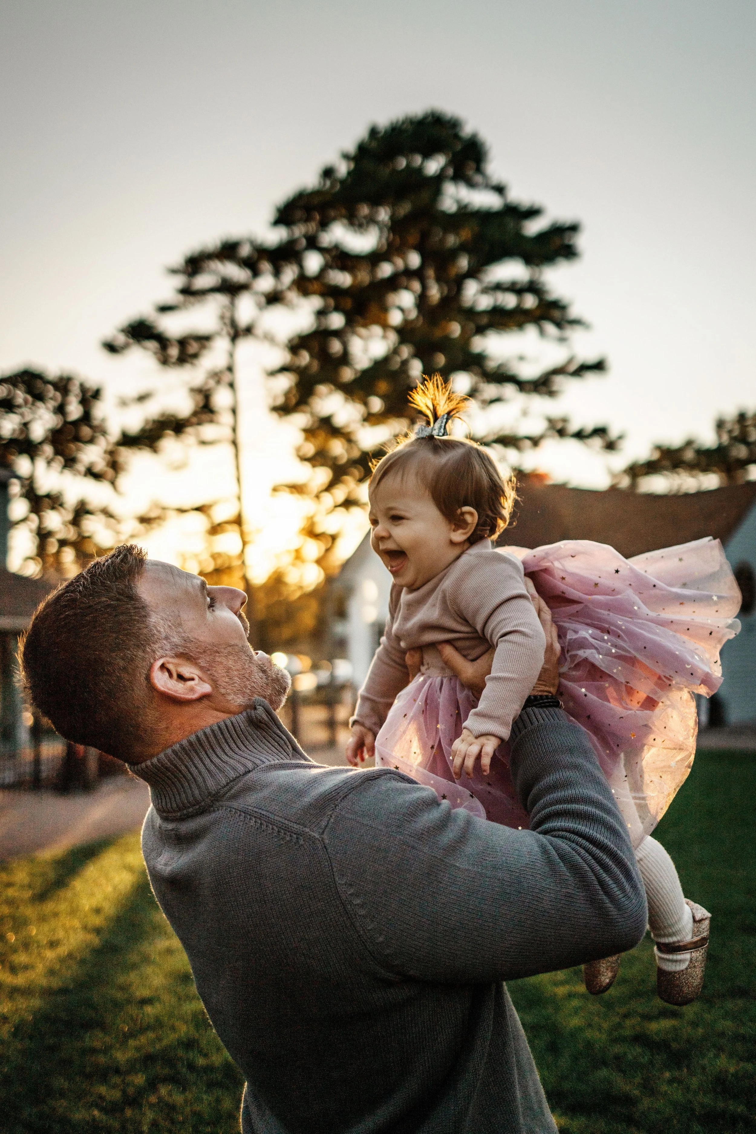 A man lifting a young girl up in his arms outdoors during sunset, with trees in the background.