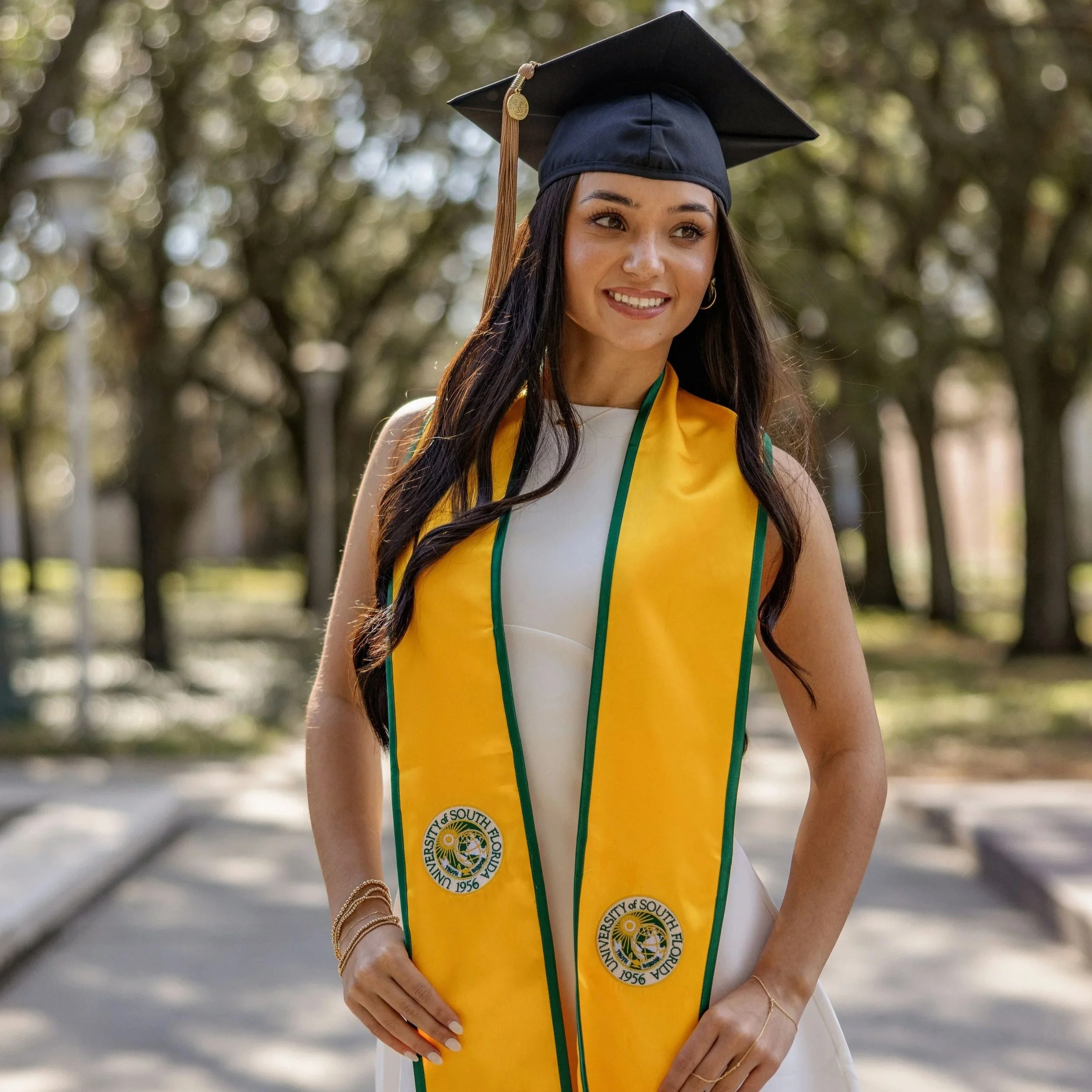 A young woman wearing a black graduation cap and a yellow stole with the University of South Florida logo, outdoors with a blurred background of trees, smiling.