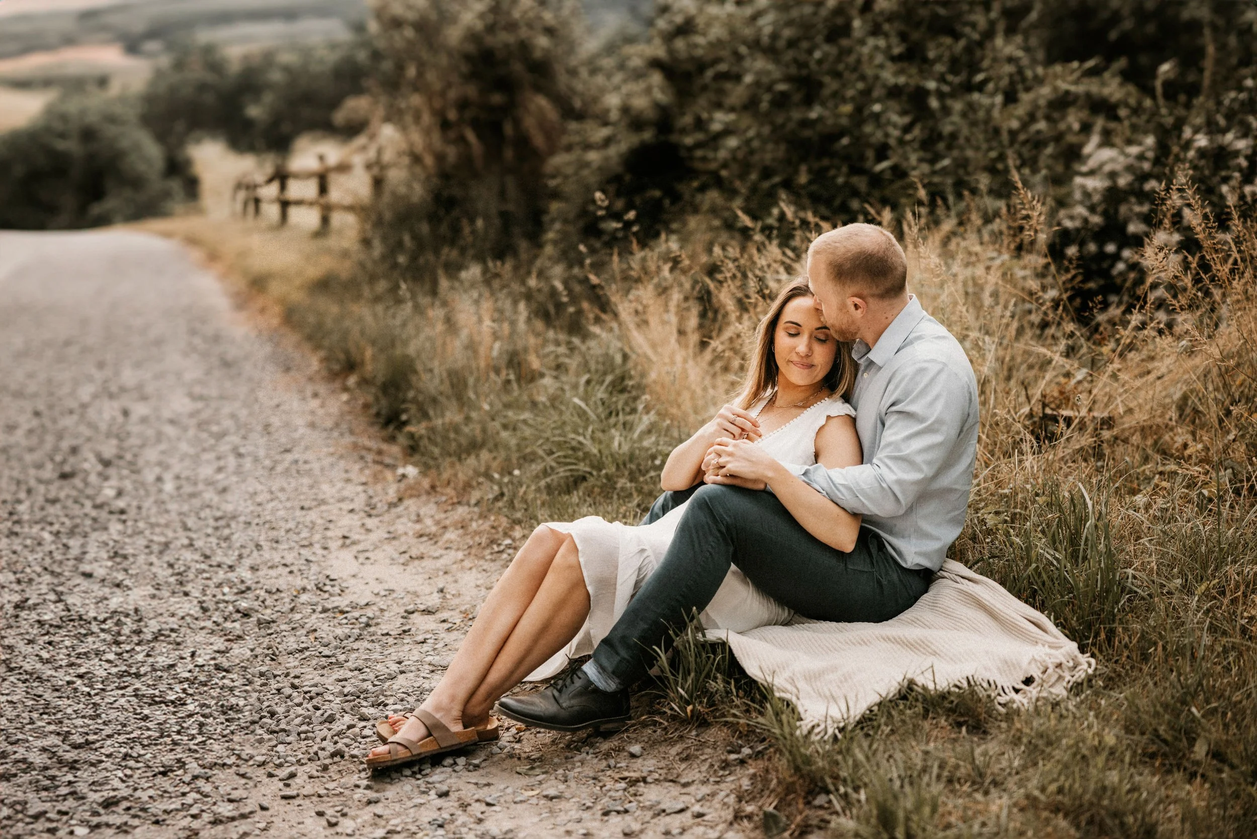 A young man and woman sit close together on a blanket beside a gravel path, with the man gently kissing the woman's forehead as she smiles peacefully. They are surrounded by tall grass and bushes, with a rustic wooden fence and countryside in the background during late afternoon or early evening.