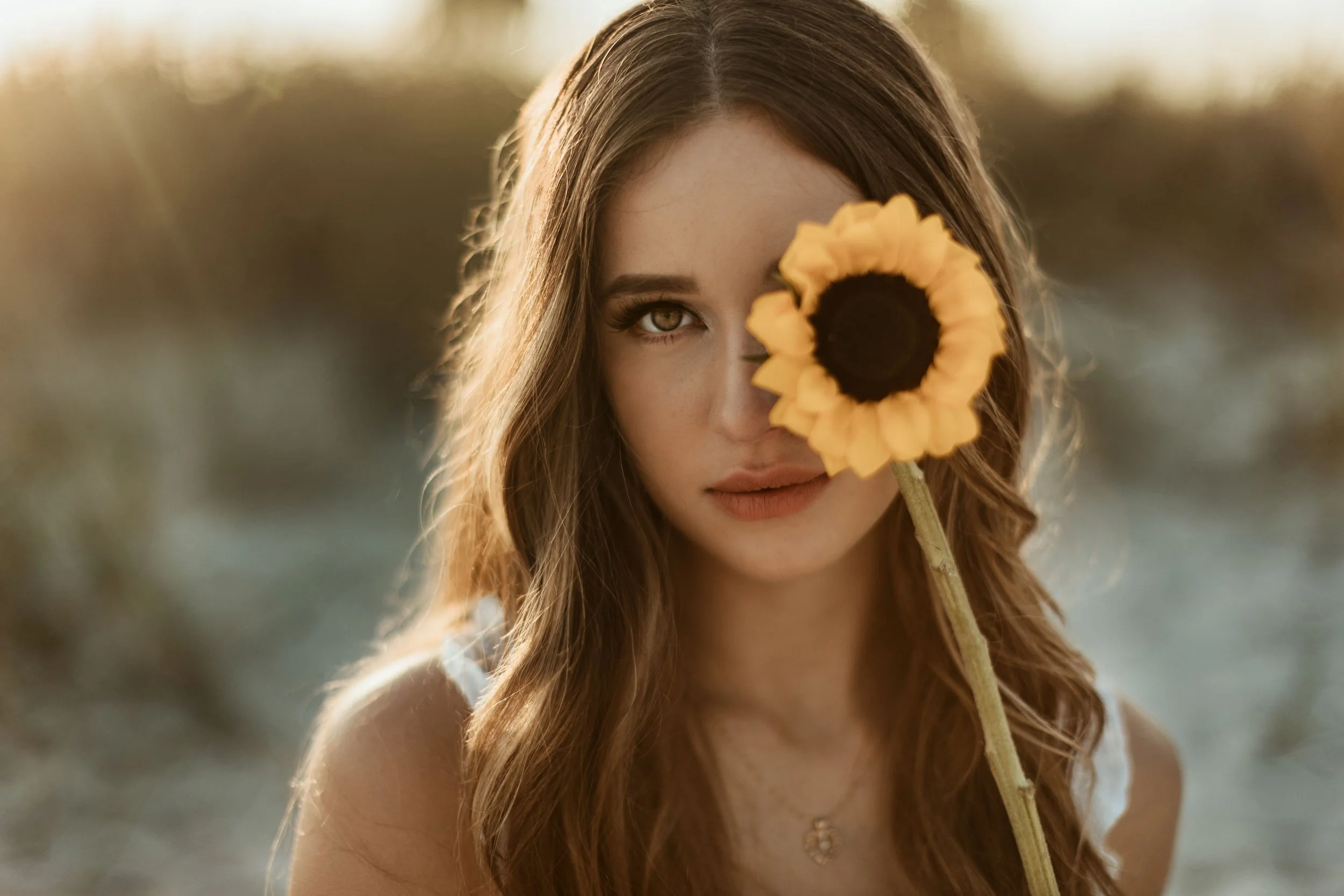 A young woman with long, wavy brown hair holding a sunflower in front of her face, covering one eye, with soft sunlight in the background.